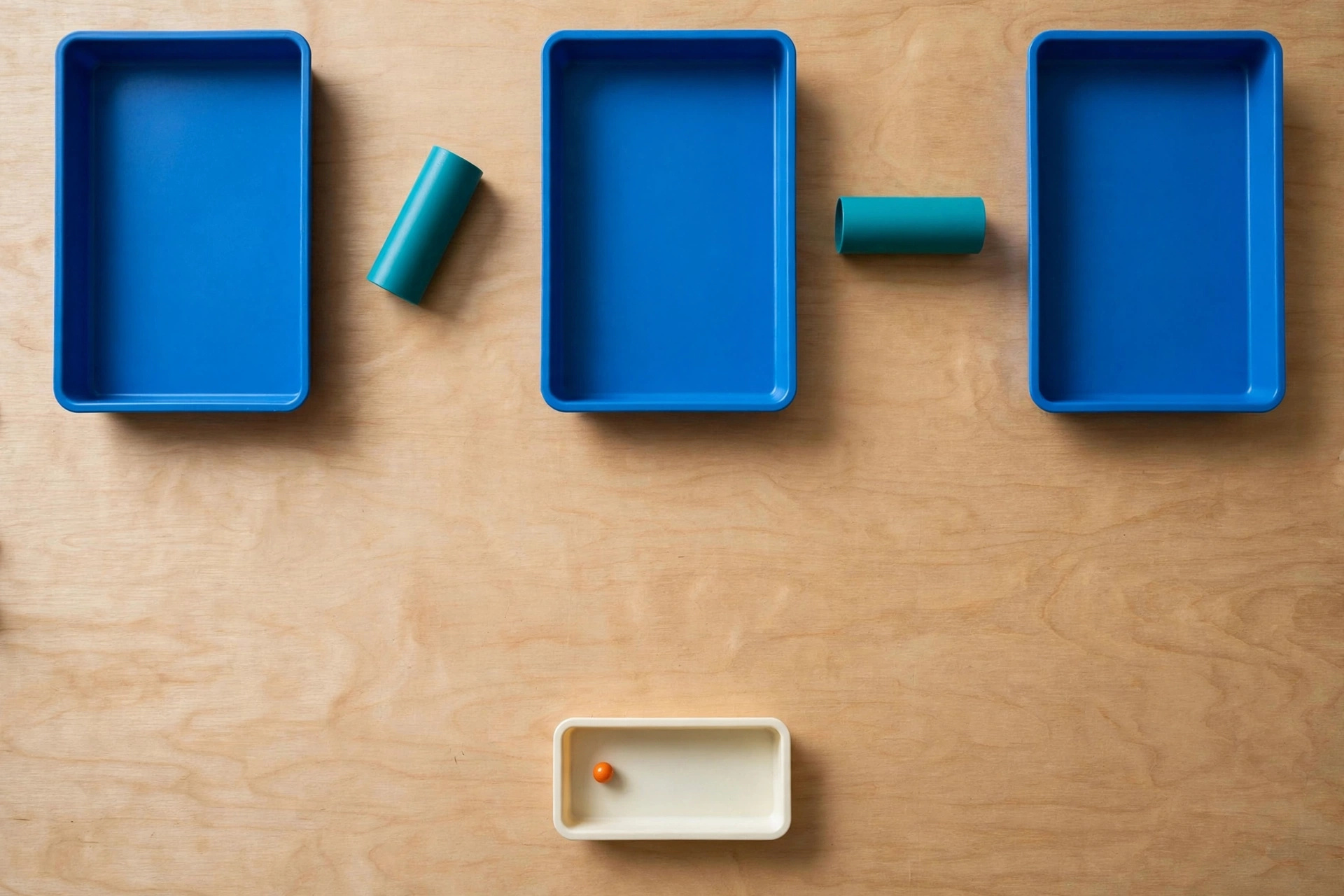 Three blue trays and two teal cylinders are arranged on a wooden surface, with a smaller ivory tray containing an orange bead below.