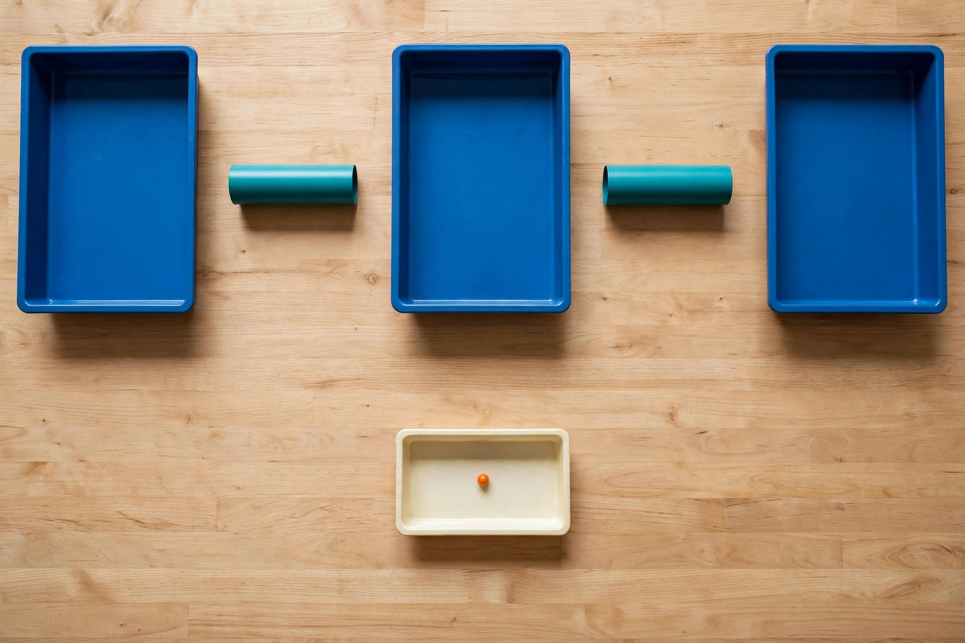 Three blue trays and two teal cylinders are arranged on a wooden surface, with a small orange bead in a separate ivory tray below.