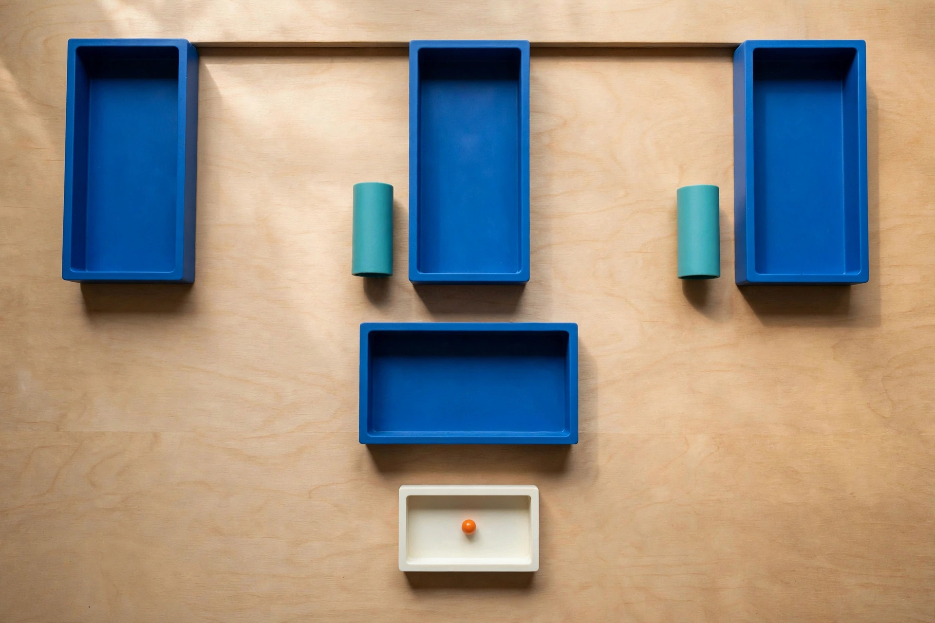 Three blue trays and two teal cylinders are arranged on a wooden surface, with a smaller ivory tray and orange bead below.