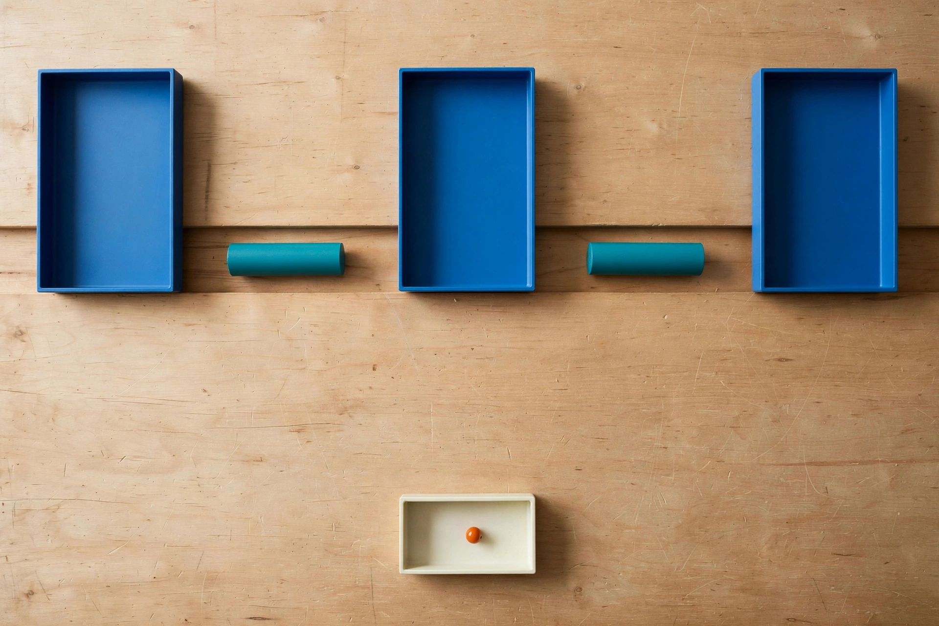 Three blue trays and two teal cylinders are arranged on a wooden surface, with a small ivory tray containing an orange bead below.