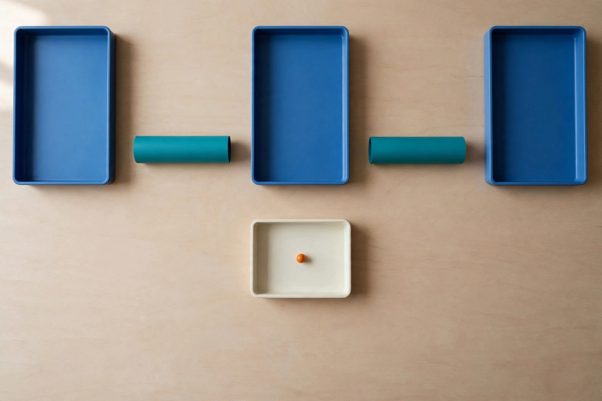 Three blue trays and two teal cylinders are arranged on a light wooden surface, with an ivory tray holding an orange bead below.