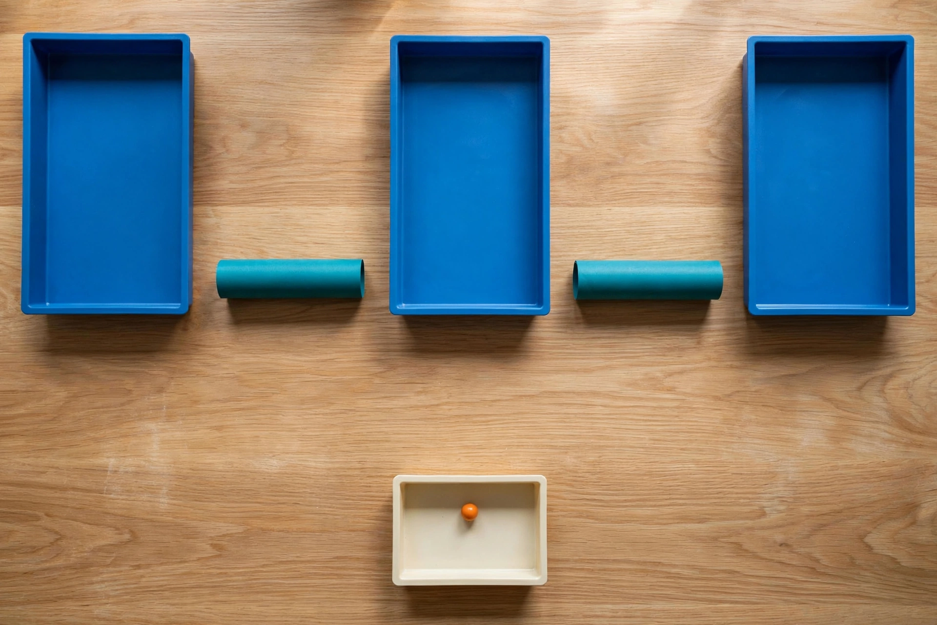 Three blue trays and two teal cylinders are arranged on a wooden surface, with a small orange bead in a separate ivory tray below.