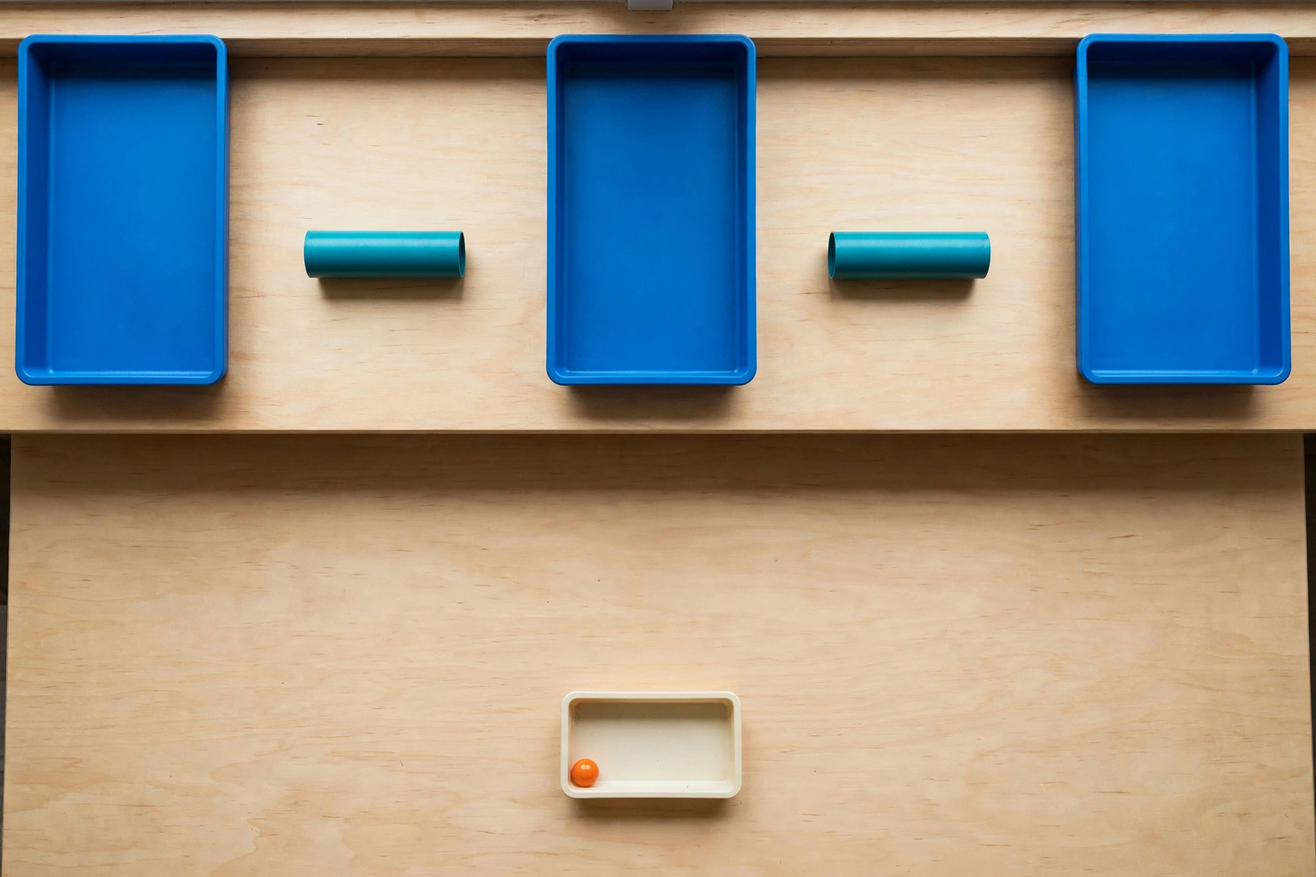 Three blue trays and two teal cylinders are arranged on a wooden surface, with an orange bead in a smaller ivory tray below.