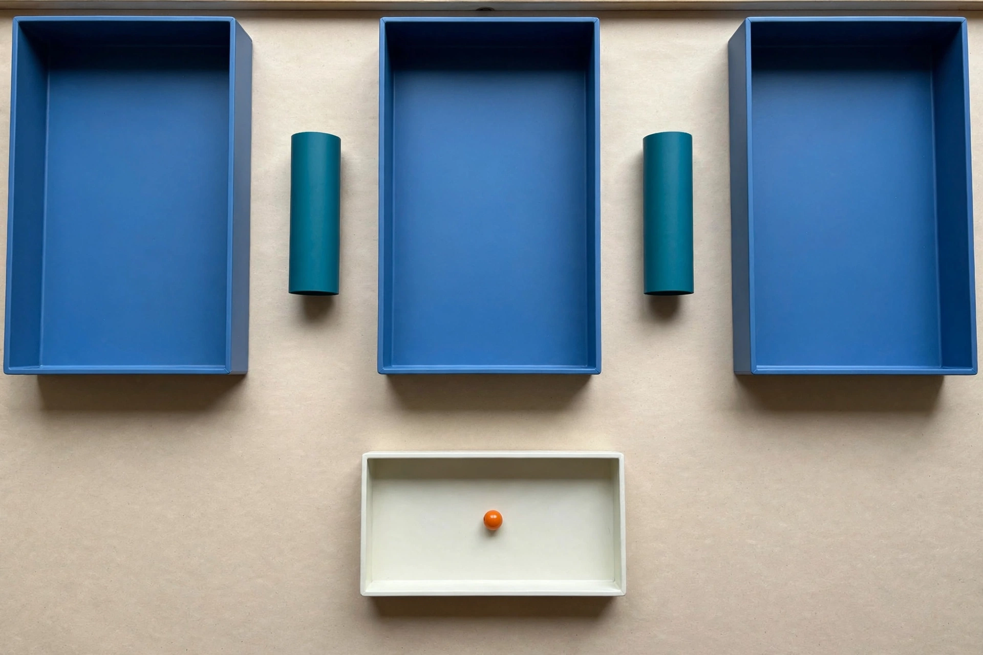 Three blue rectangular trays are arranged in a row with gaps between them, each containing a teal cylinder. Below, a single ivory tray holds an orange bead.