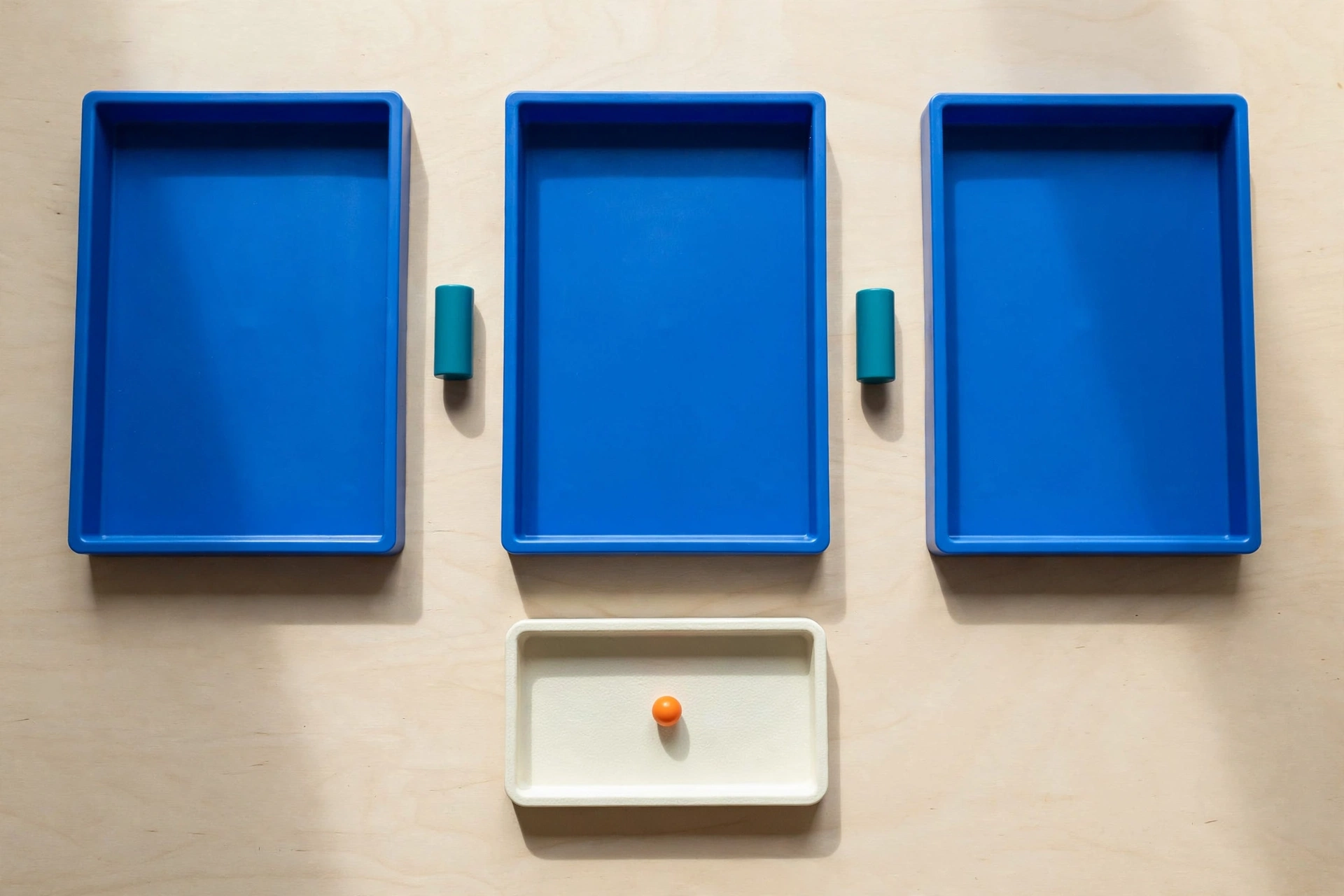 Three blue trays and two teal cylinders are arranged in a row on a wooden surface, with an ivory tray and orange bead below.