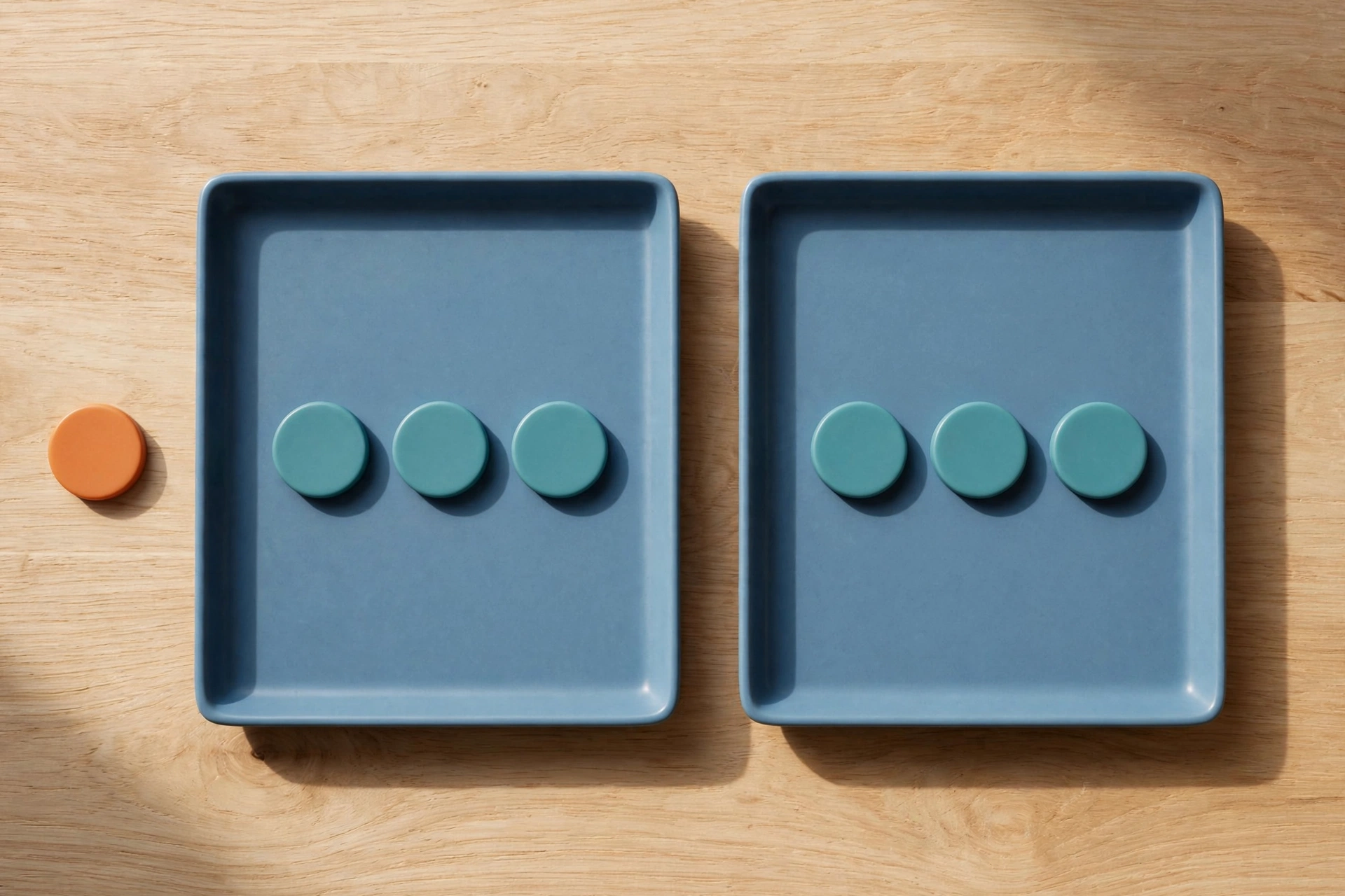 Two blue trays with teal tokens sit side-by-side on a wooden surface, with an orange token placed beside the left tray.