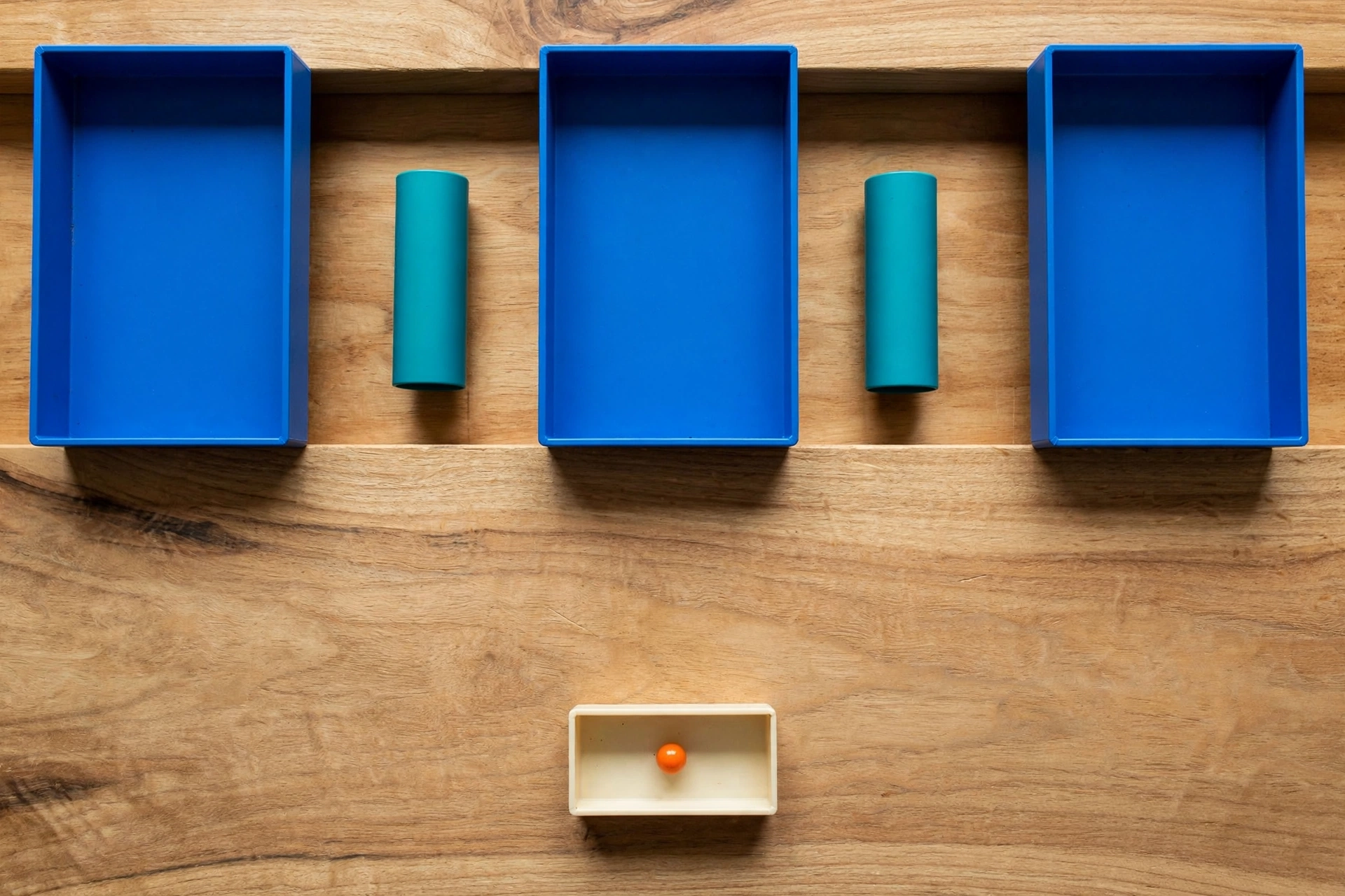 Three blue trays and two teal cylinders are arranged on a wooden surface, with an orange bead in a separate ivory tray below.