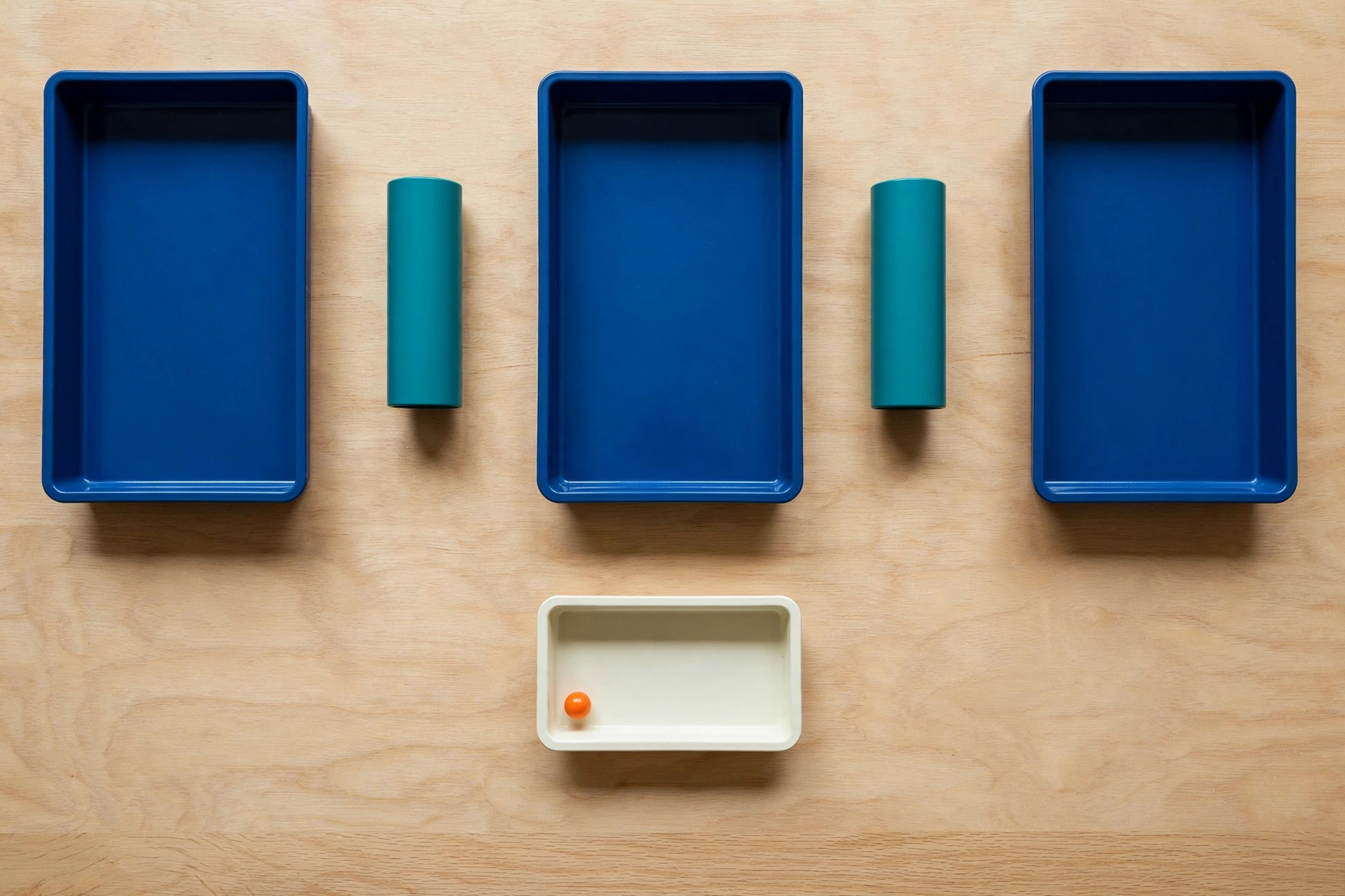 Three blue trays and two teal cylinders are arranged on a wooden surface, with an orange bead in a separate ivory tray below.