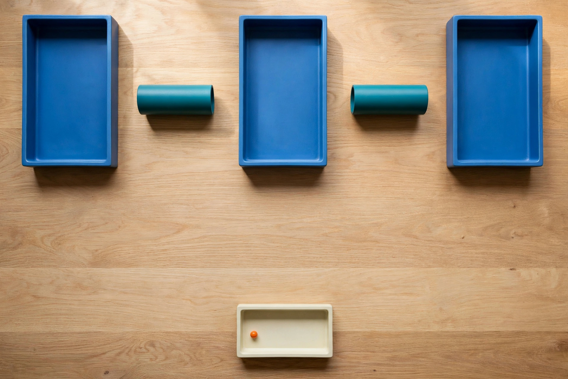 Three blue rectangular trays and two teal cylinders are arranged on a wooden desk, with a smaller ivory tray containing an orange bead below.