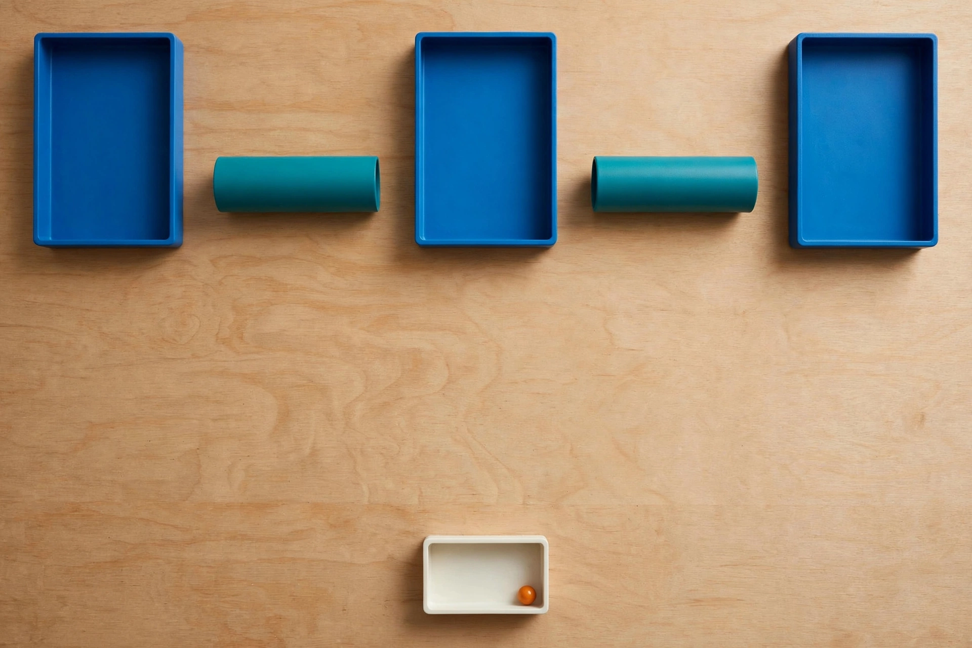 Three blue trays and two teal cylinders are arranged on a wooden surface, with a small ivory tray holding an orange bead below.