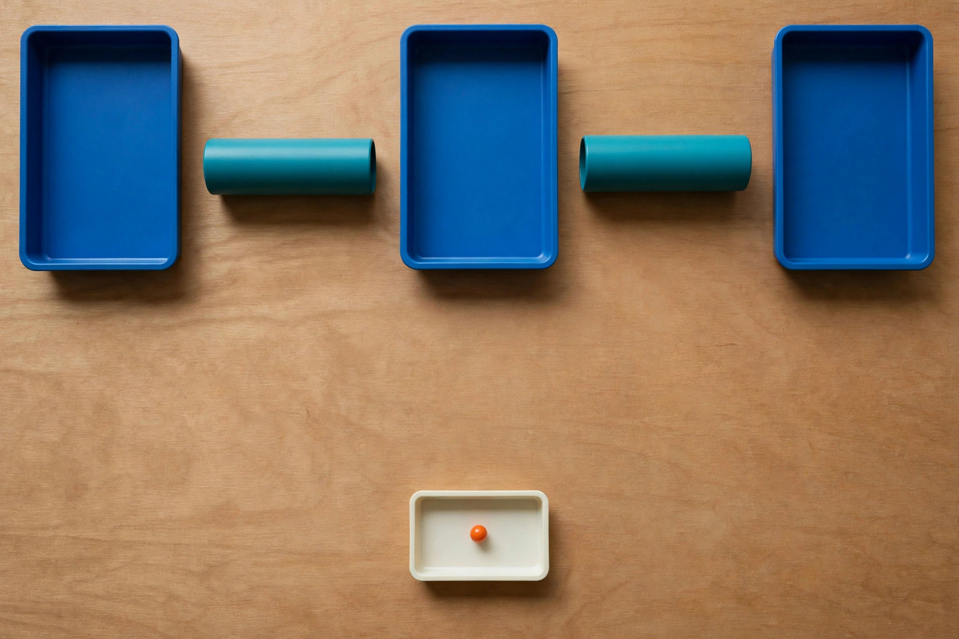 Three blue trays and two teal cylinders are arranged on a wooden surface, with a smaller ivory tray containing an orange bead below.