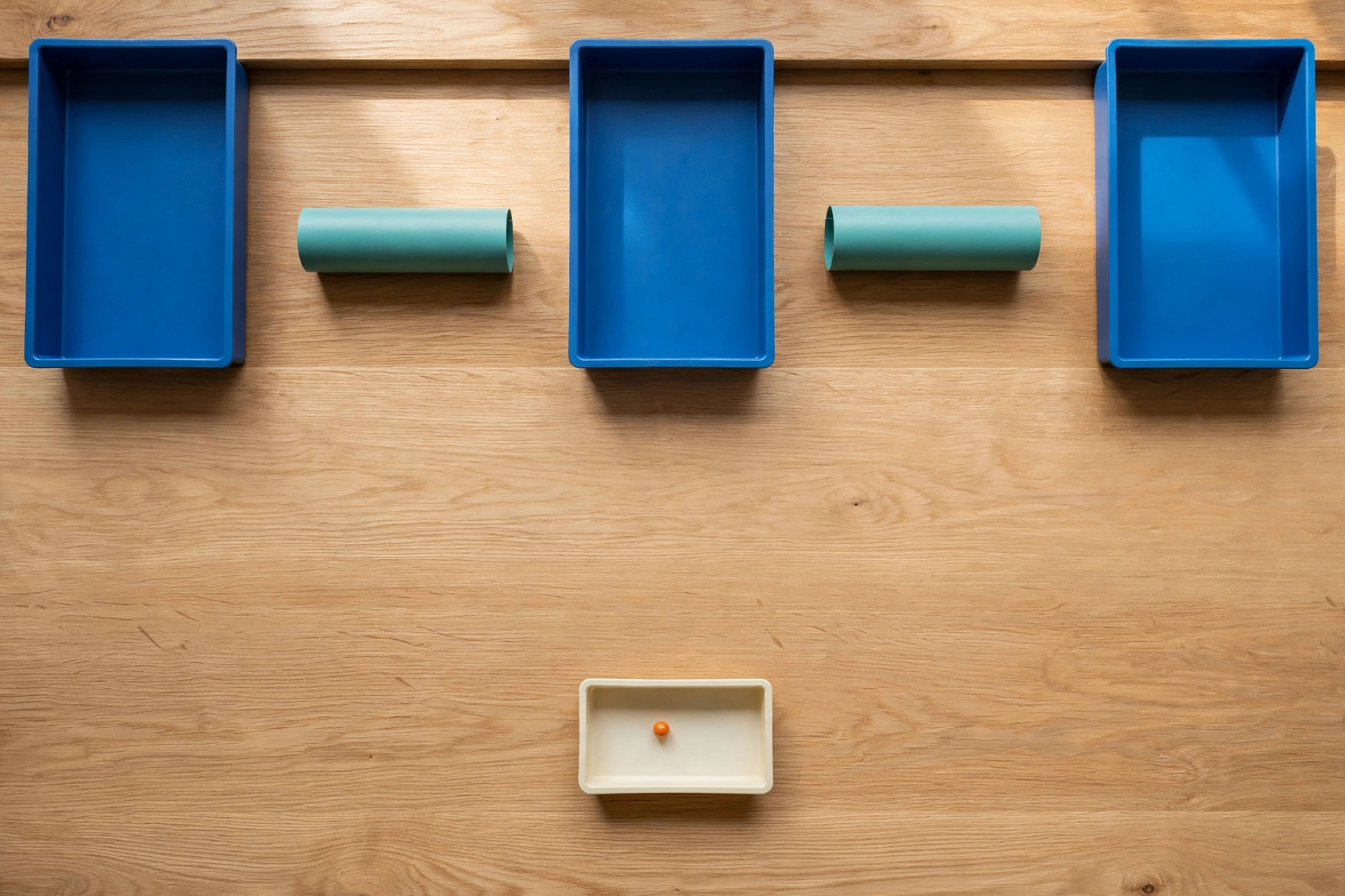 Three blue trays and two teal cylinders are arranged on a wooden surface, with a small orange bead in a lower ivory tray.