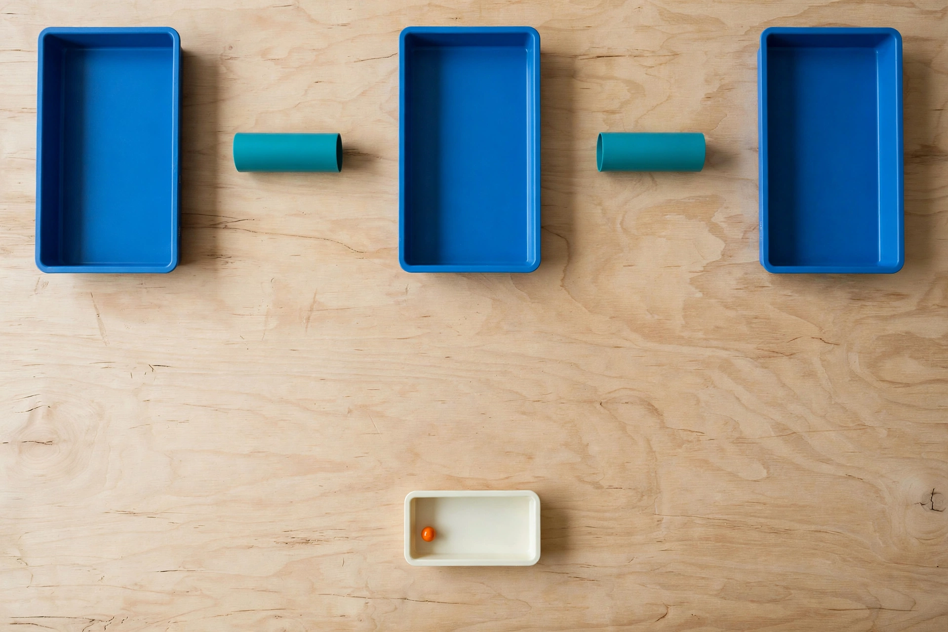 Three blue trays and two teal cylinders are arranged on a wooden surface, with a small ivory tray holding an orange bead below.
