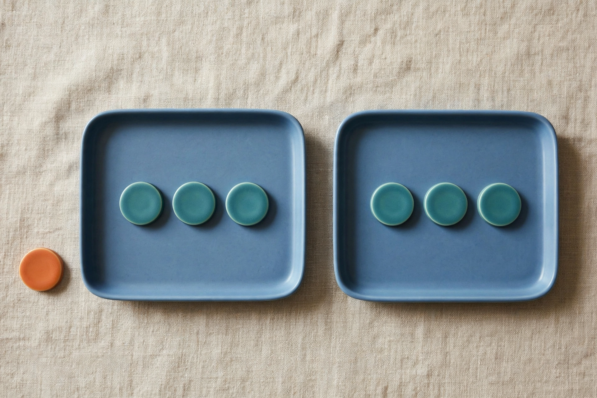 Two blue trays with three teal tokens each sit on a textured surface, with one orange token placed beside the left tray.