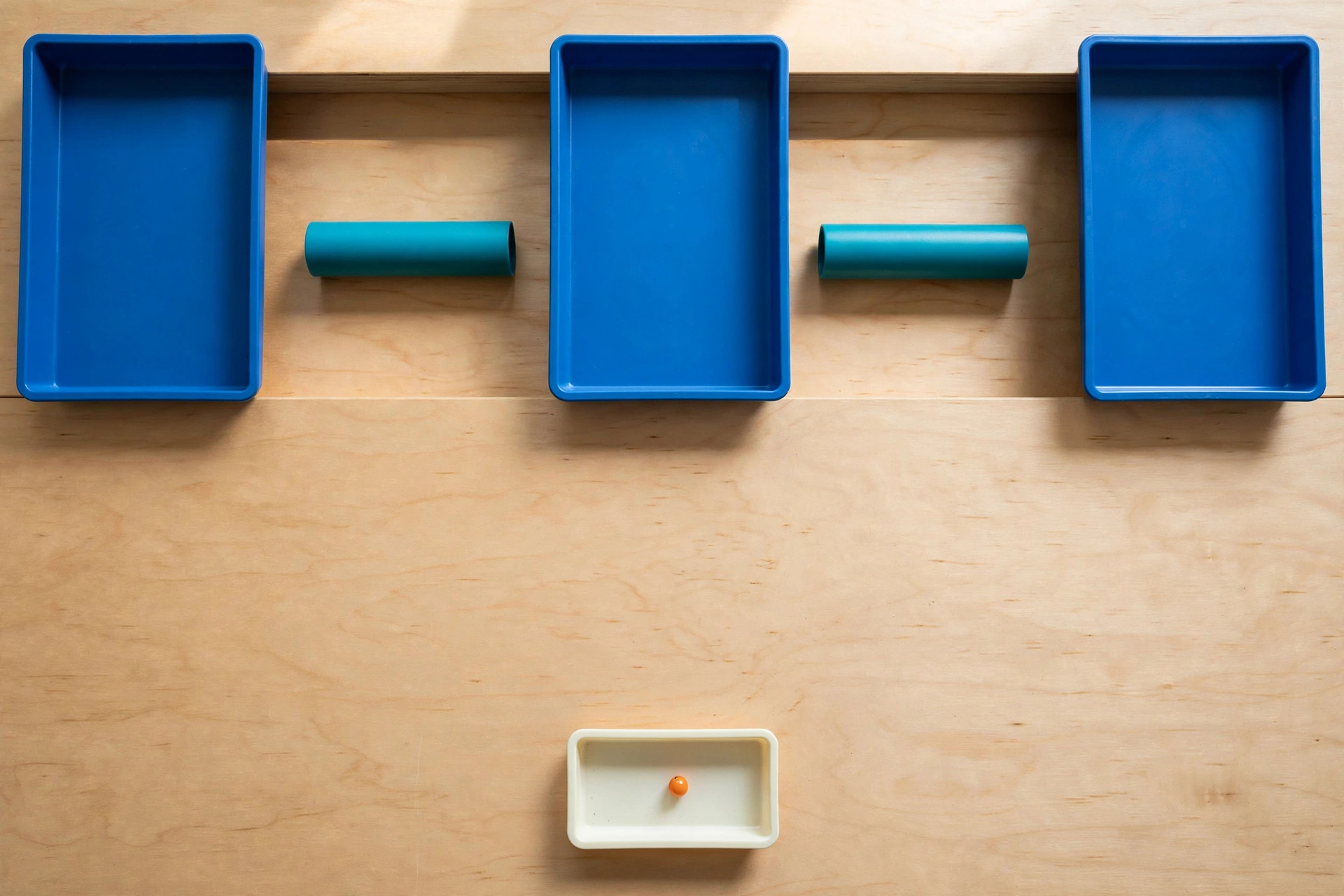 Three blue trays and two teal cylinders are arranged on a wooden surface, with a small orange bead in a separate ivory tray below.