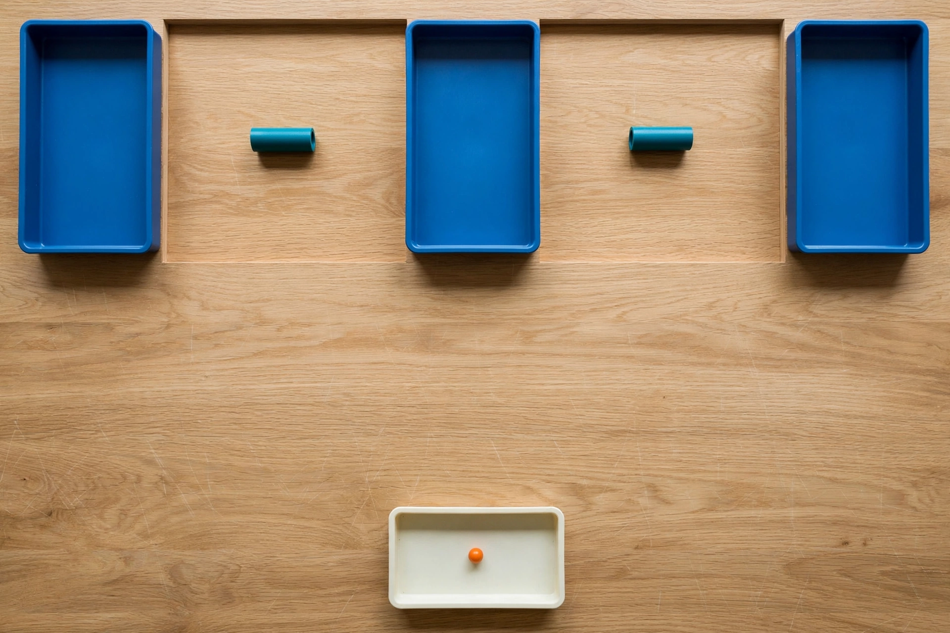 Three blue trays and two teal cylinders are arranged on a wooden surface, with a small ivory tray containing an orange bead below.