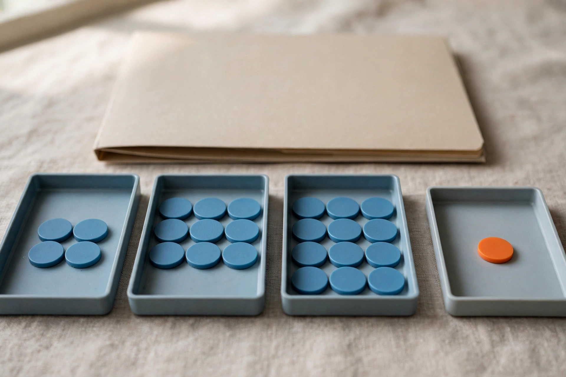 Four shallow office trays with blue tokens progress from left to right, with a fourth tray holding a single orange token, and a folder behind them.