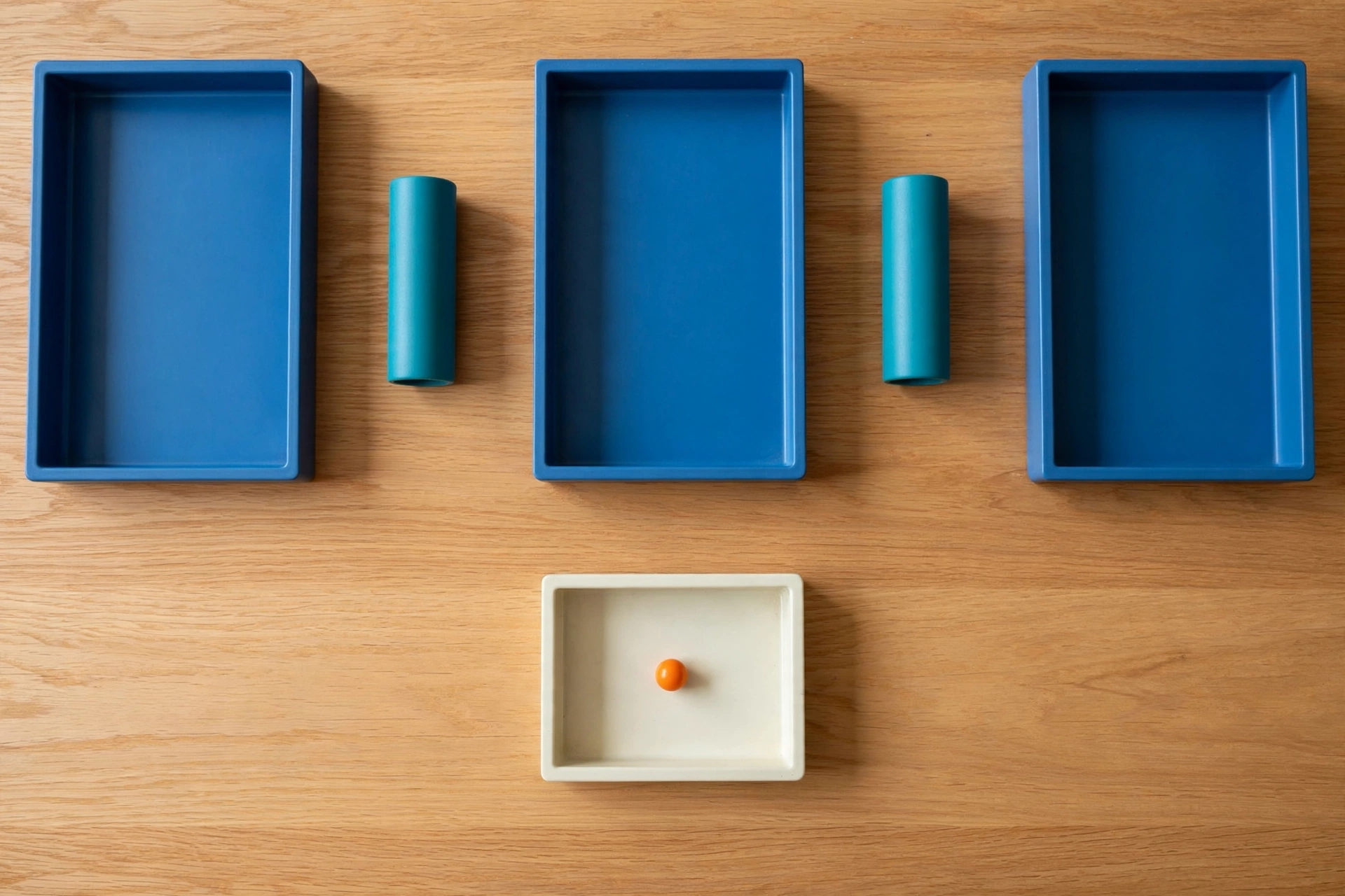 Three blue trays and two teal cylinders are arranged on a wooden surface, with one orange bead in a lower ivory tray.