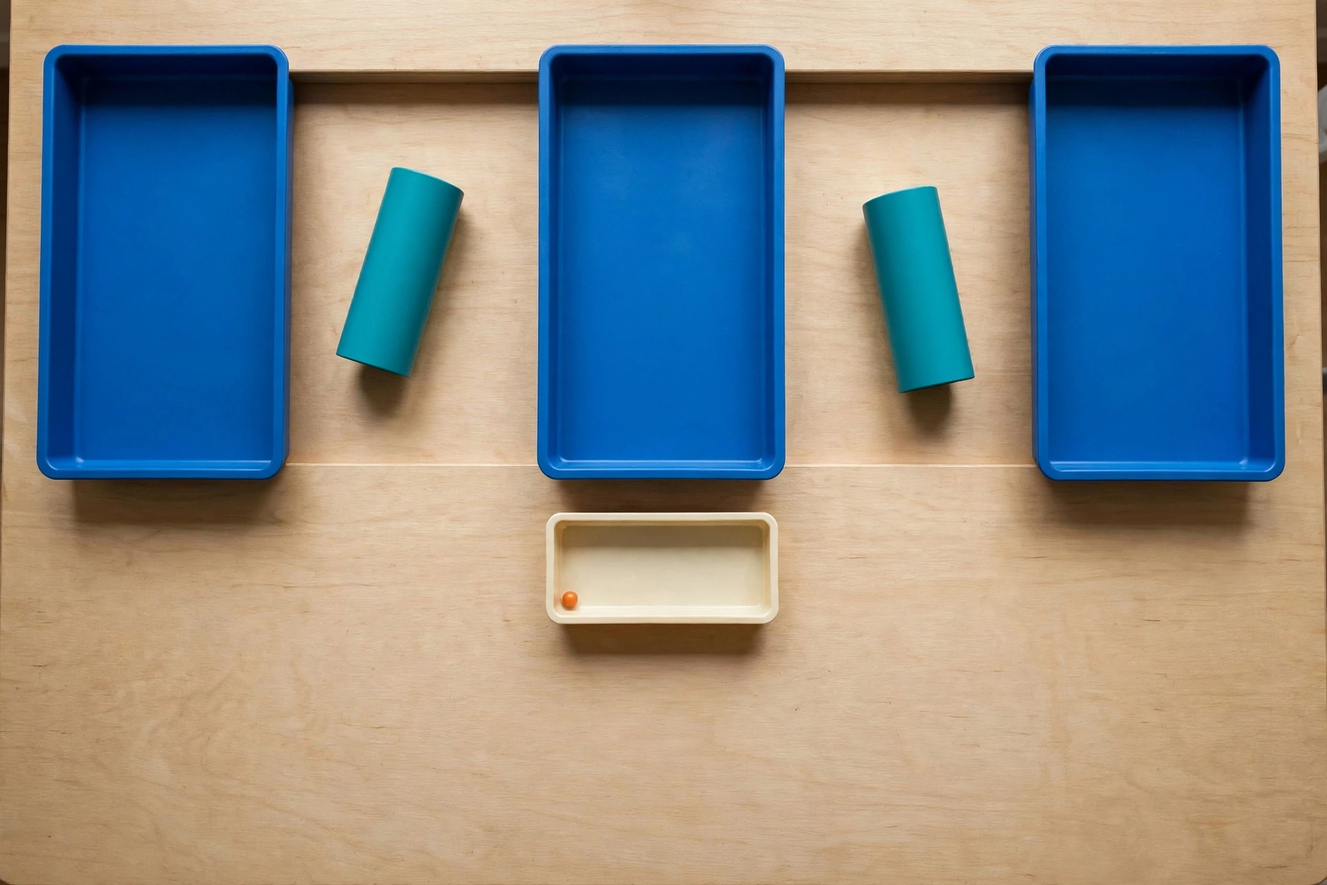 Three blue trays and two teal cylinders are arranged on a wooden surface, with an orange bead in a small ivory tray below.