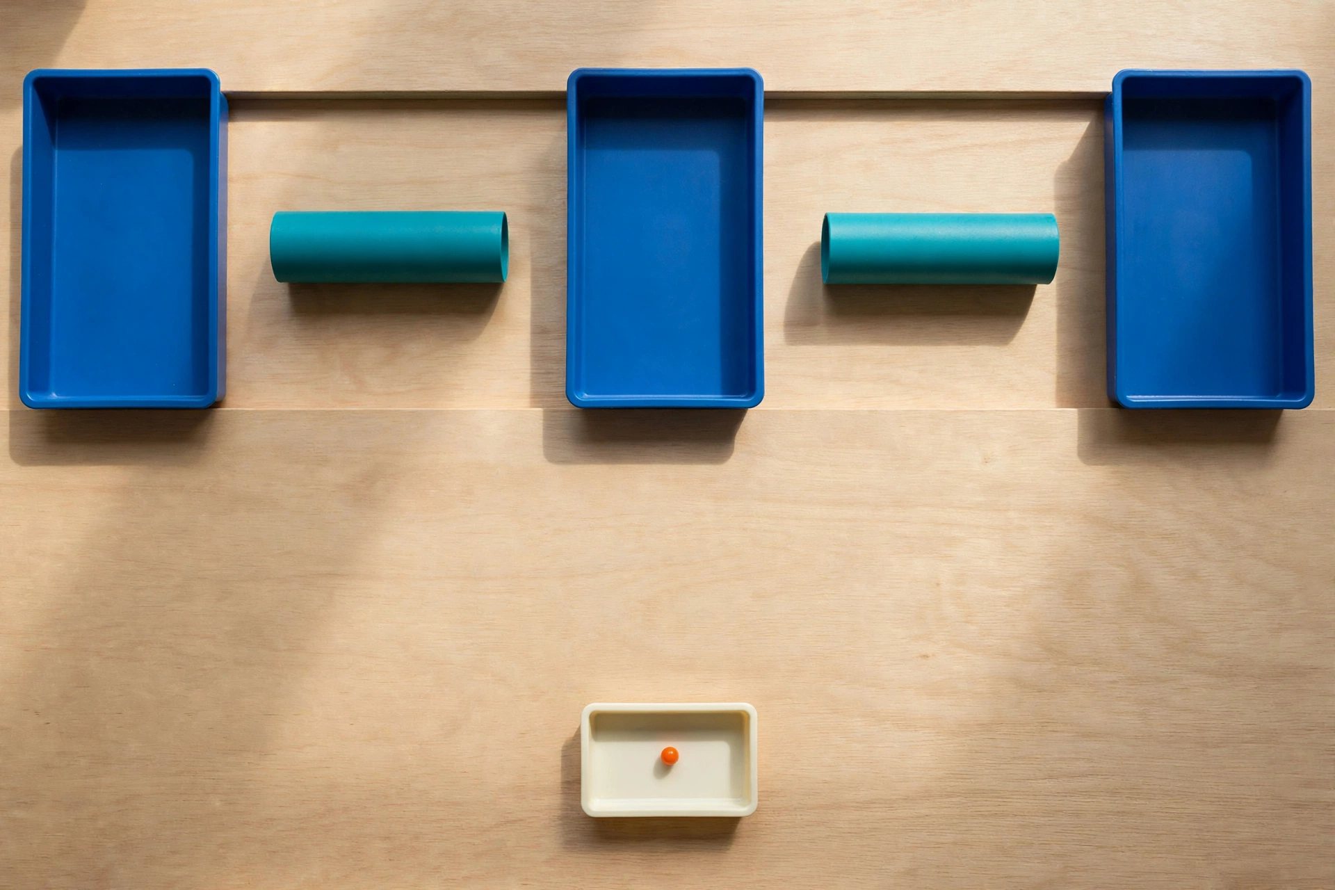 Three blue trays and two teal cylinders are arranged in a row on a wooden surface, with a smaller ivory tray containing an orange bead below.