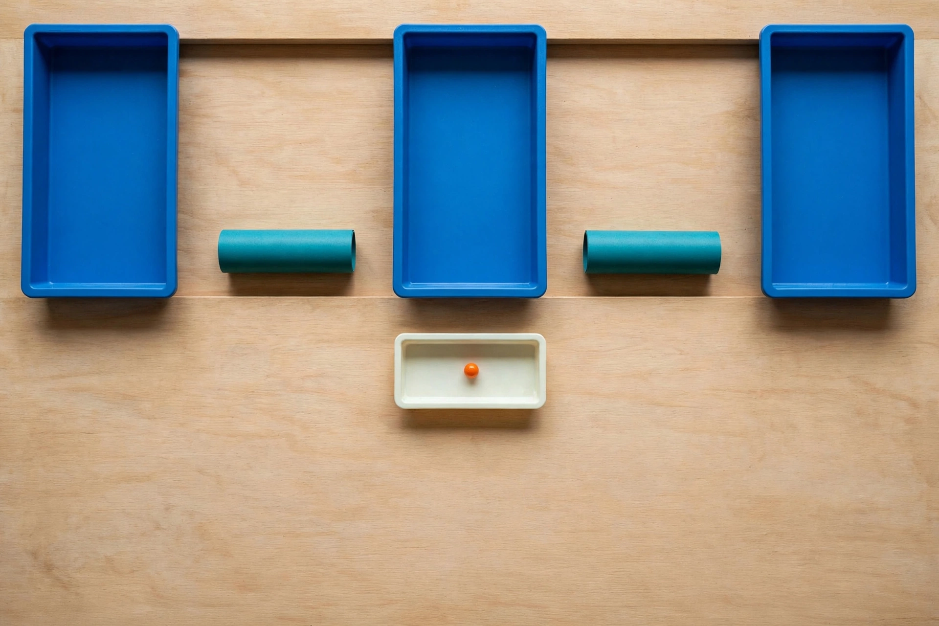 Three blue trays and two teal cylinders are arranged on a wooden surface, with an ivory tray containing an orange bead below.