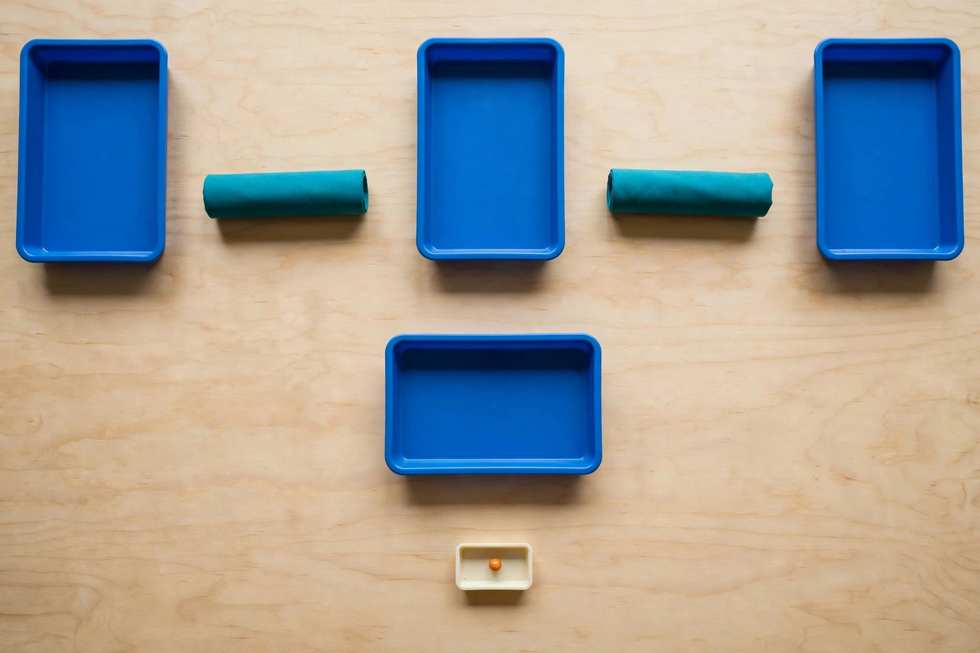 Three blue trays are arranged in a row with two teal cylinders in the gaps between them, and a small ivory tray with an orange bead below.