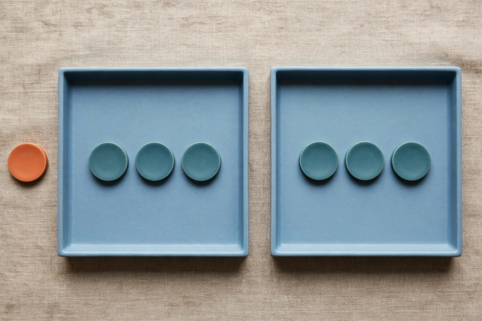 Two blue trays each hold three teal tokens, with one orange token placed beside the left tray on a textured surface.