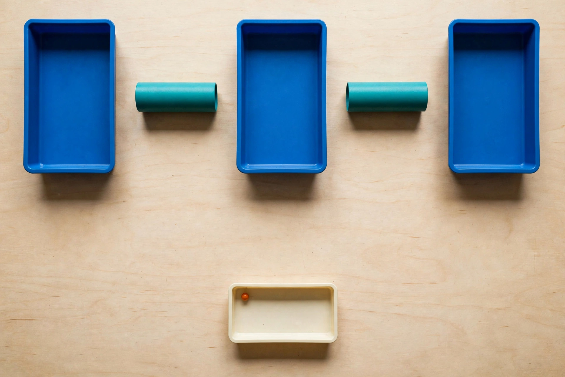 Three blue trays are arranged in a row with gaps between them, each containing a teal cylinder. A fourth ivory tray sits below with an orange bead.