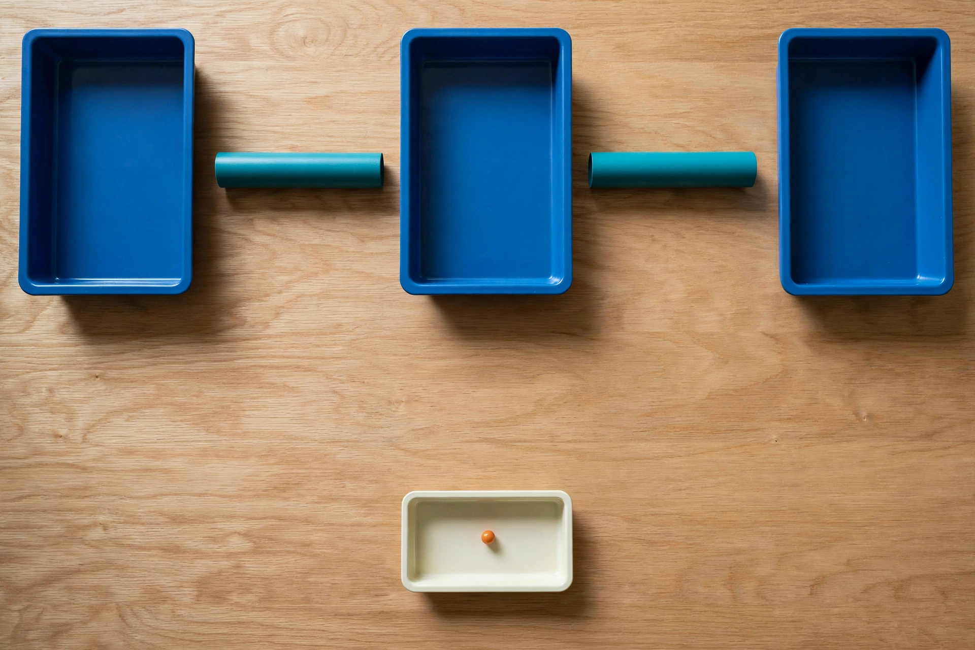 Three blue trays are arranged in a row with teal cylinders between them, and an ivory tray with an orange bead sits below.