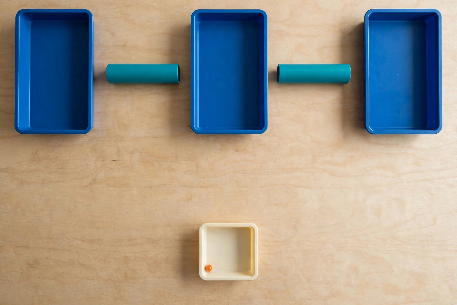Three blue trays and two teal cylinders are arranged on a wooden surface, with a smaller ivory tray containing an orange bead below.