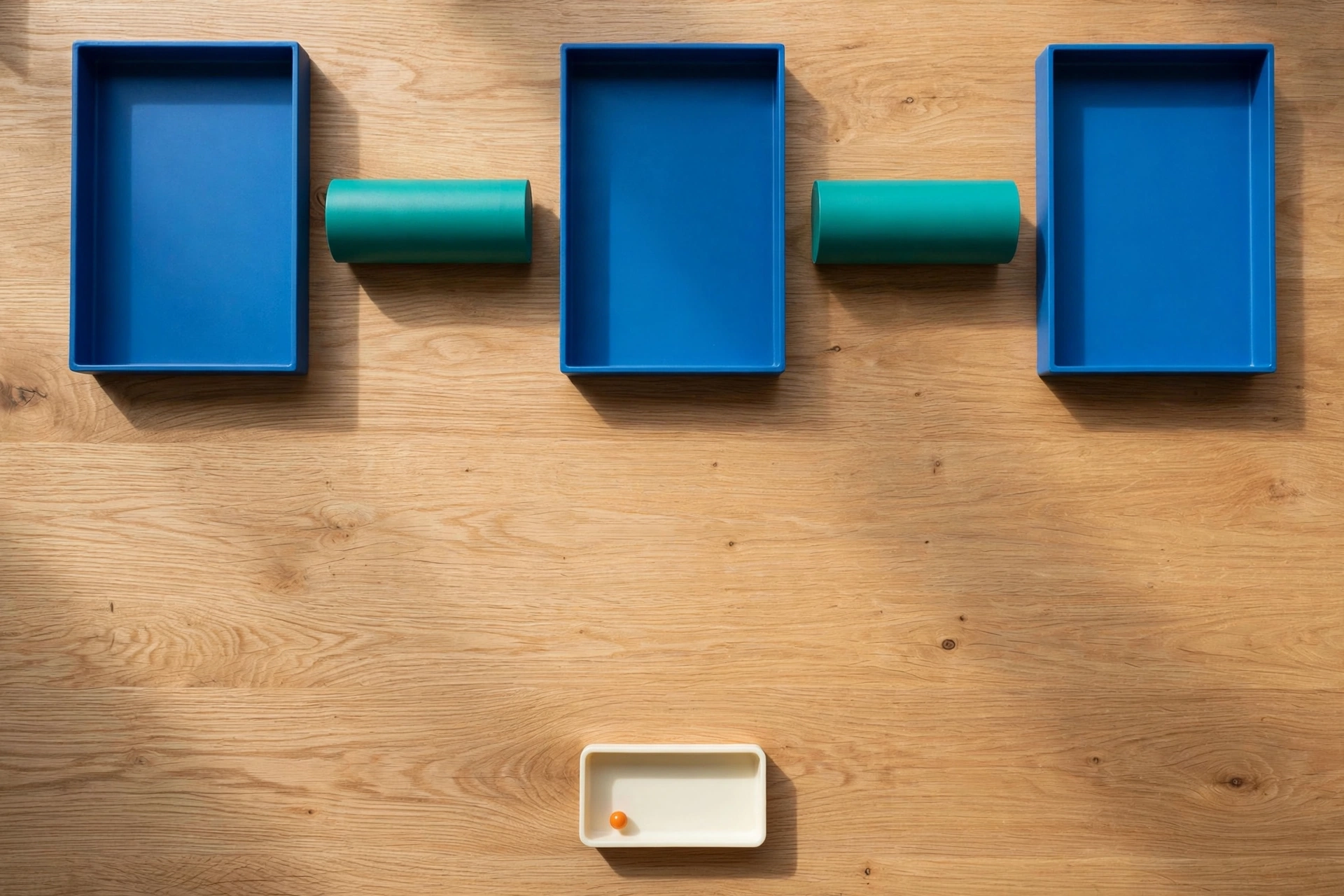 Three blue trays and two teal cylinders are arranged on a wooden desk, with a small ivory tray containing an orange bead below.
