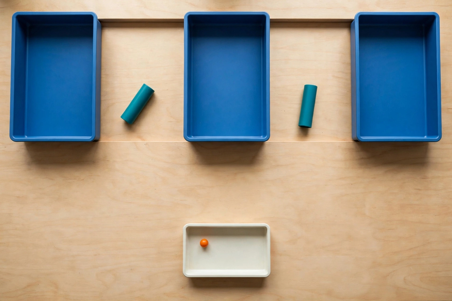 Three blue trays and two teal cylinders are arranged on a wooden surface, with a small orange bead in a separate ivory tray below.