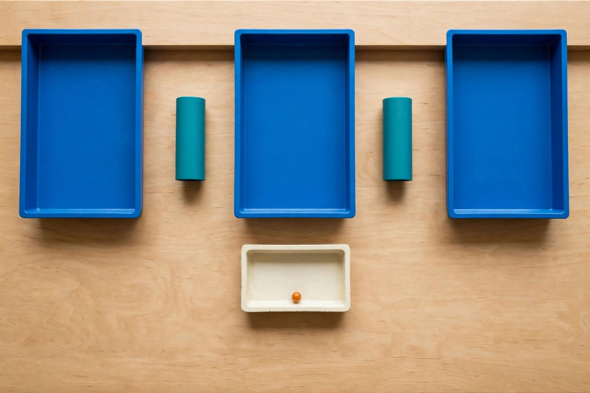 Three blue trays and two teal cylinders are arranged on a wooden surface, with an orange bead in a lower ivory tray.