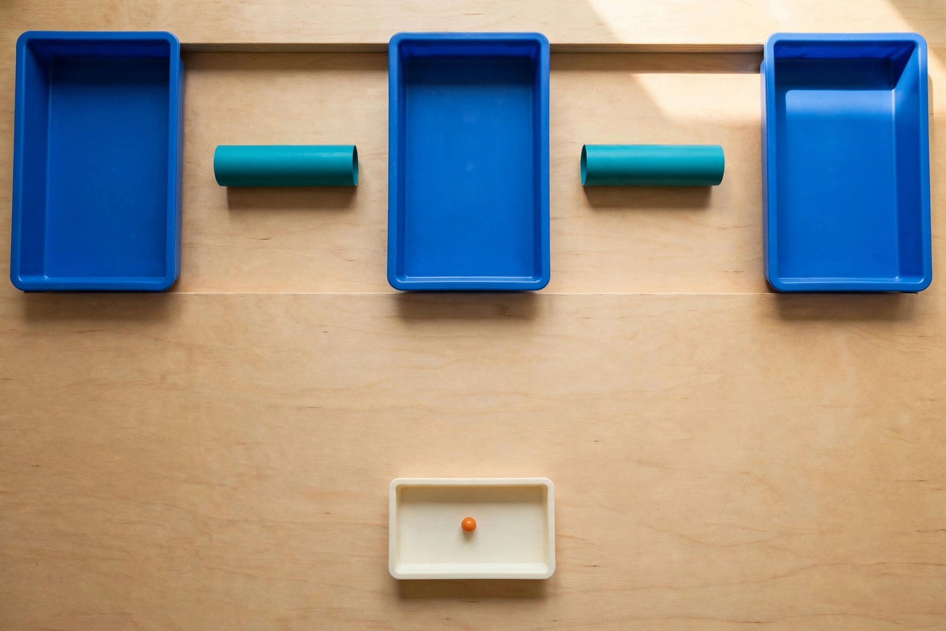 Three blue trays and two teal cylinders are arranged on a wooden surface, with an orange bead in a smaller ivory tray below.
