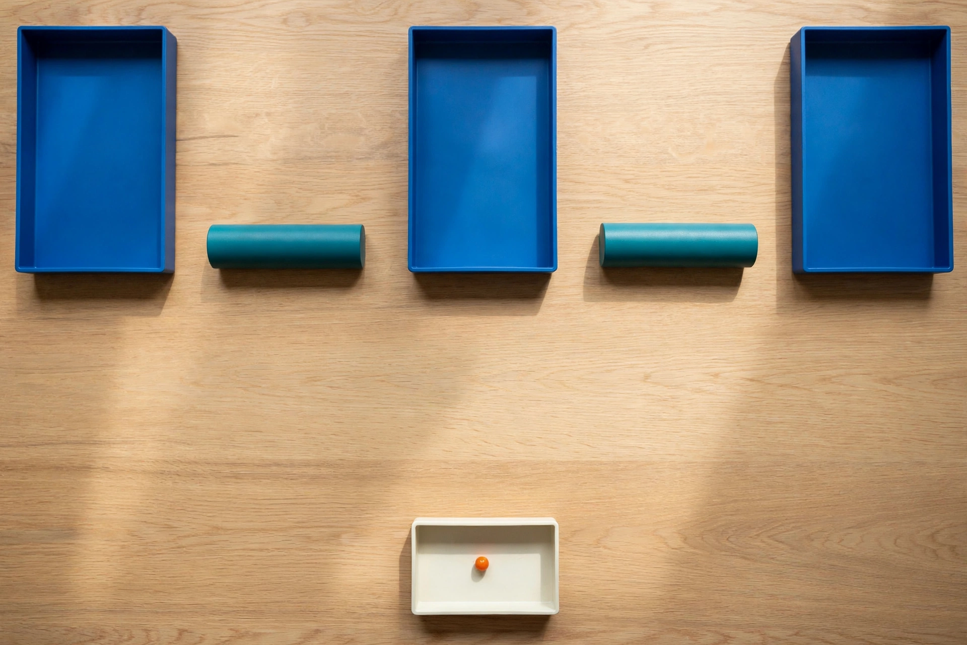 Three blue trays and two teal cylinders are arranged on a wooden surface, with a small orange bead in a white tray below.