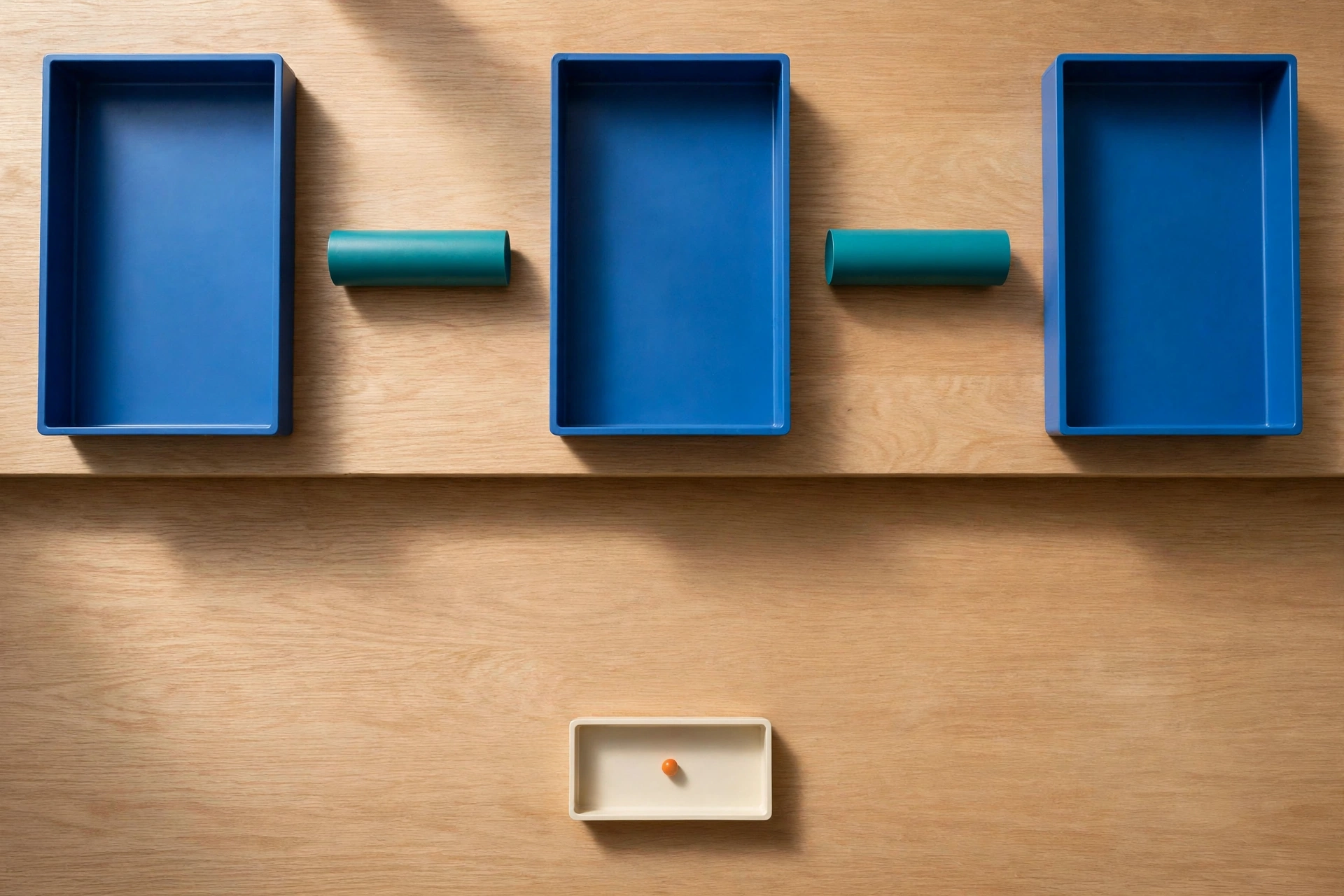 Three blue trays and two teal cylinders are arranged on a wooden surface, with a small ivory tray holding an orange bead below.