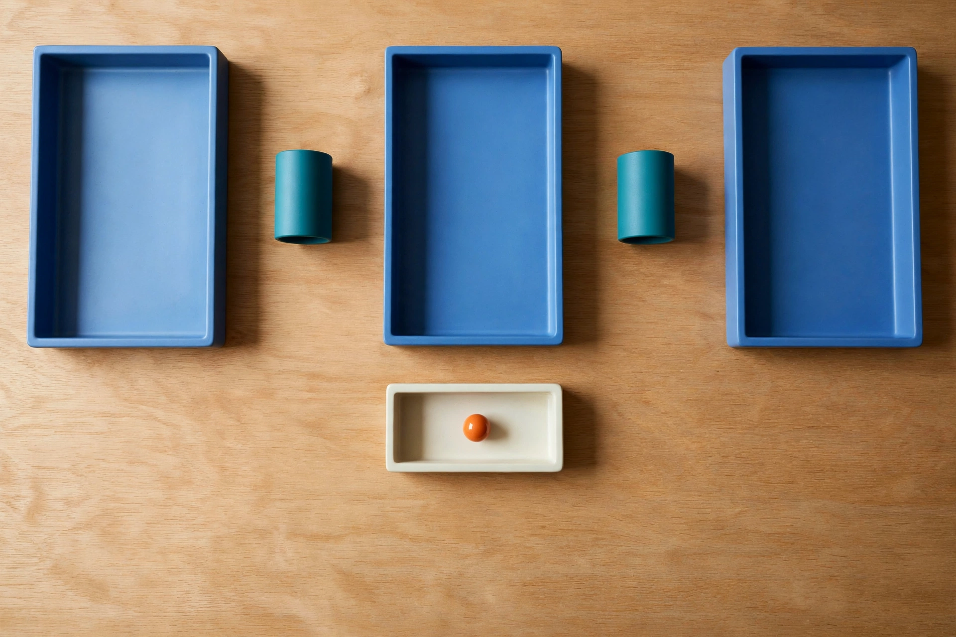 Three blue trays and two teal cylinders are arranged on a wooden surface, with an ivory tray holding an orange bead below.