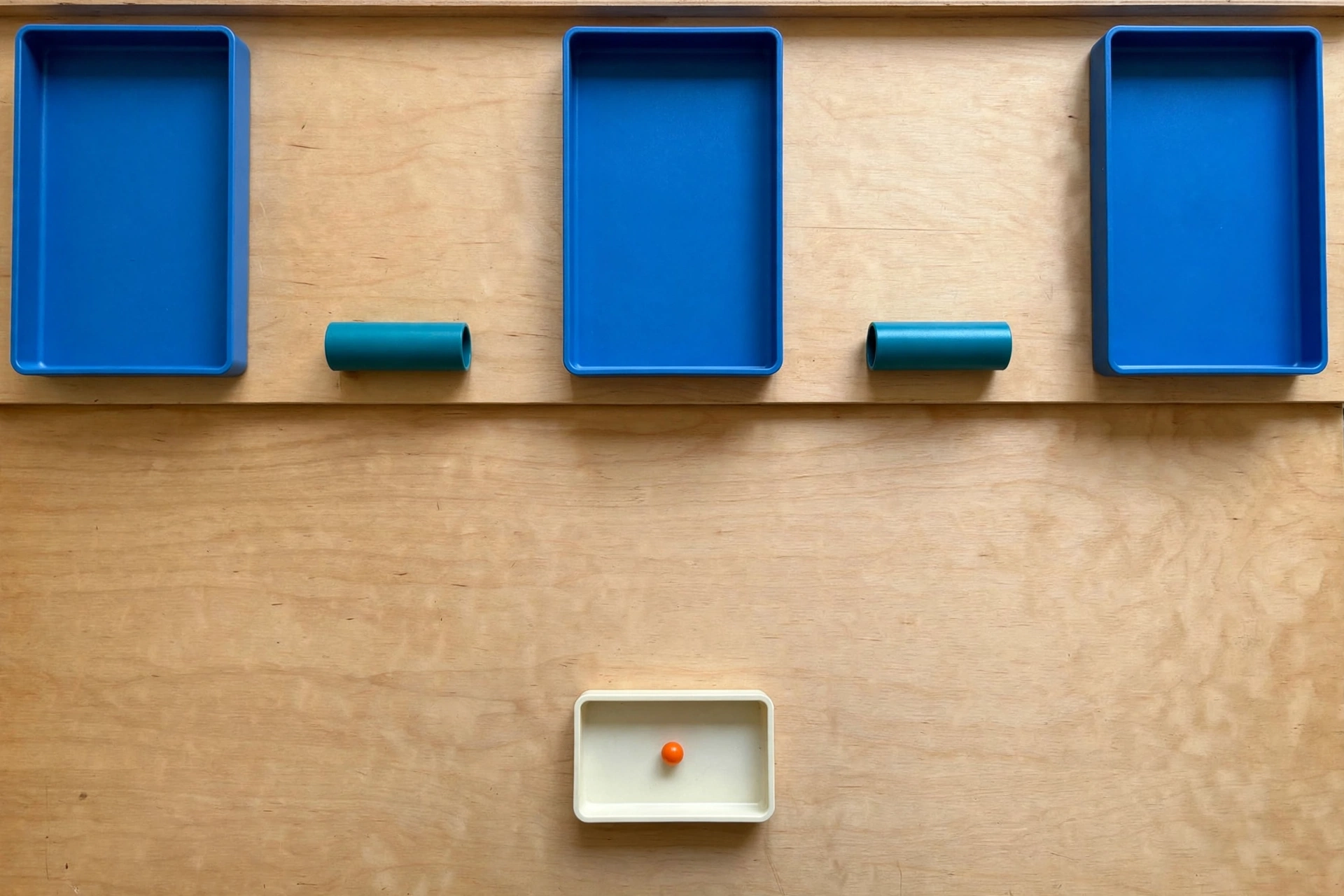 Three blue trays and two teal cylinders are arranged on a wooden surface, with an orange bead in a separate ivory tray below.