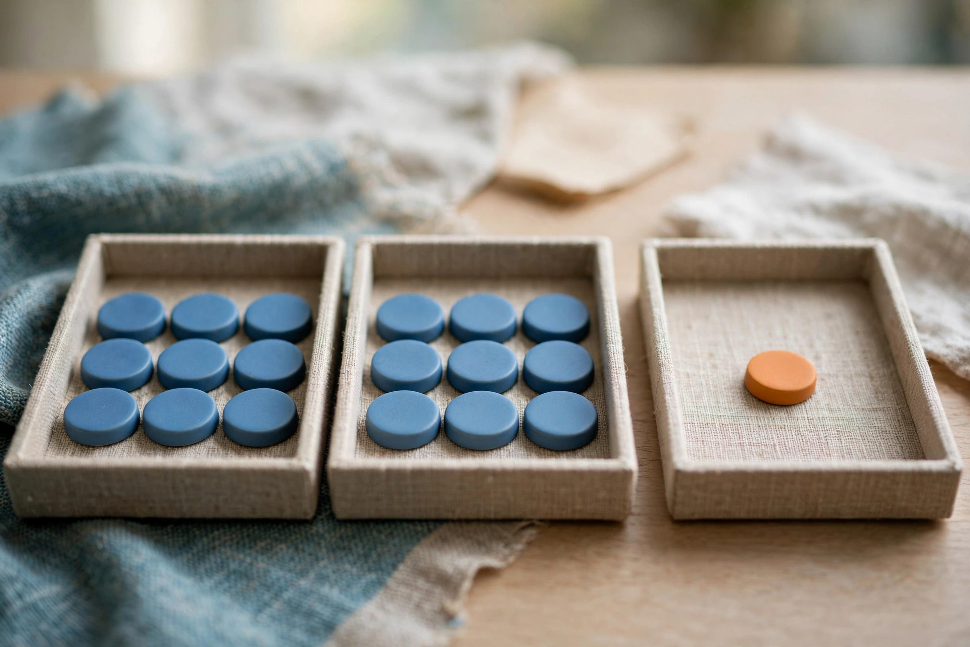 Three shallow trays of tokens sit on a wooden surface, two filled with identical blue tokens and one with a single orange token.