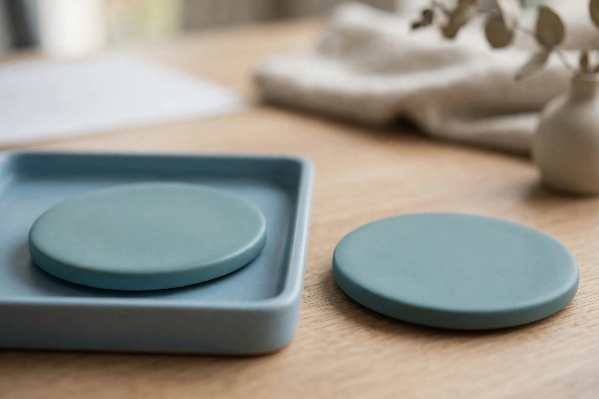 Two identical teal ceramic discs are shown on a wooden desk; one disc rests in a blue tray, and the other is placed separately beside it.