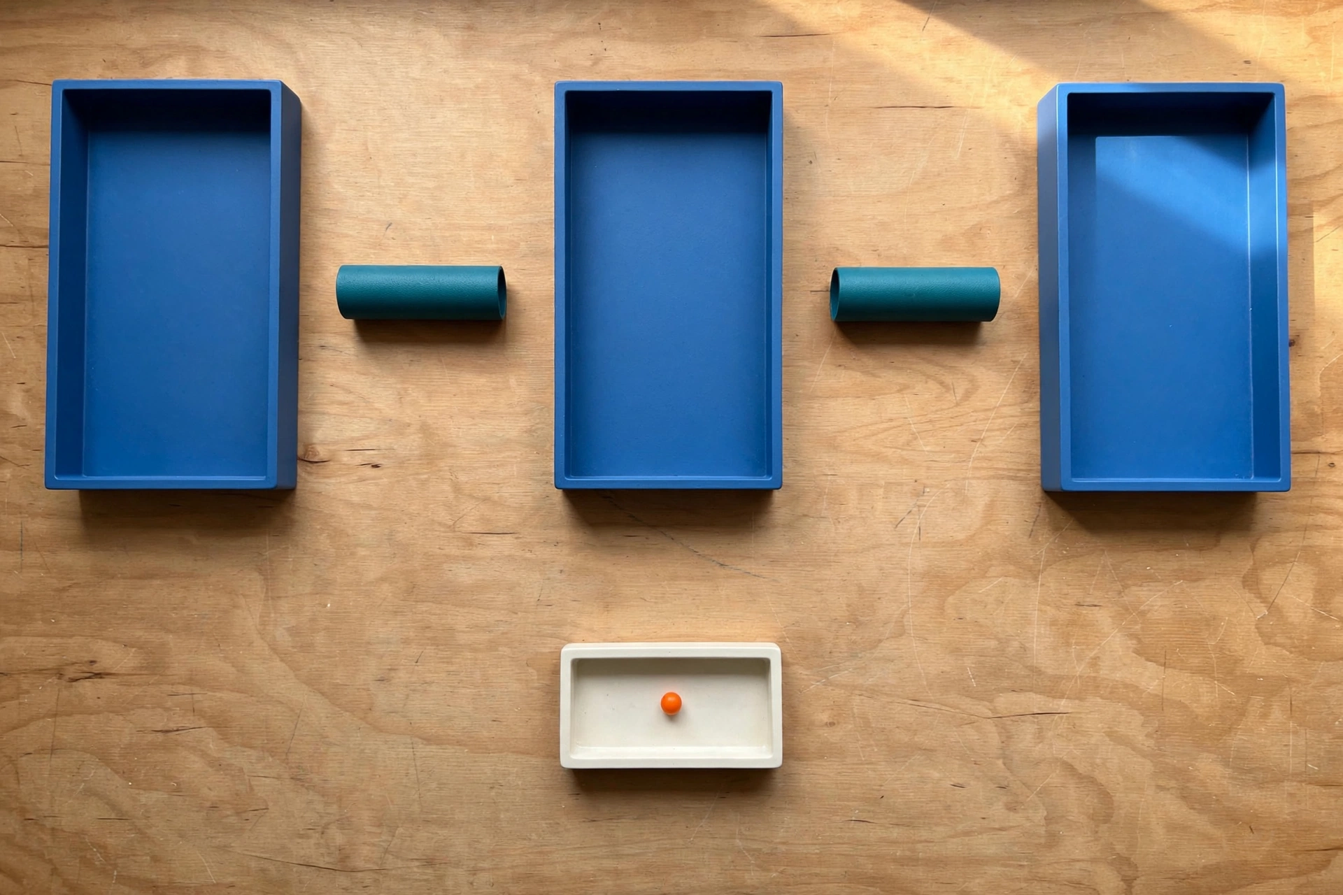 Three blue trays and two teal cylinders are arranged on a wooden surface, with a small ivory tray containing an orange bead below.