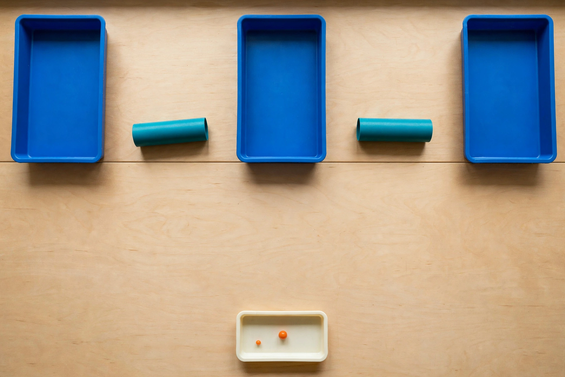 Three blue trays and two teal cylinders are arranged on a wooden surface, with a smaller ivory tray containing two orange beads below.