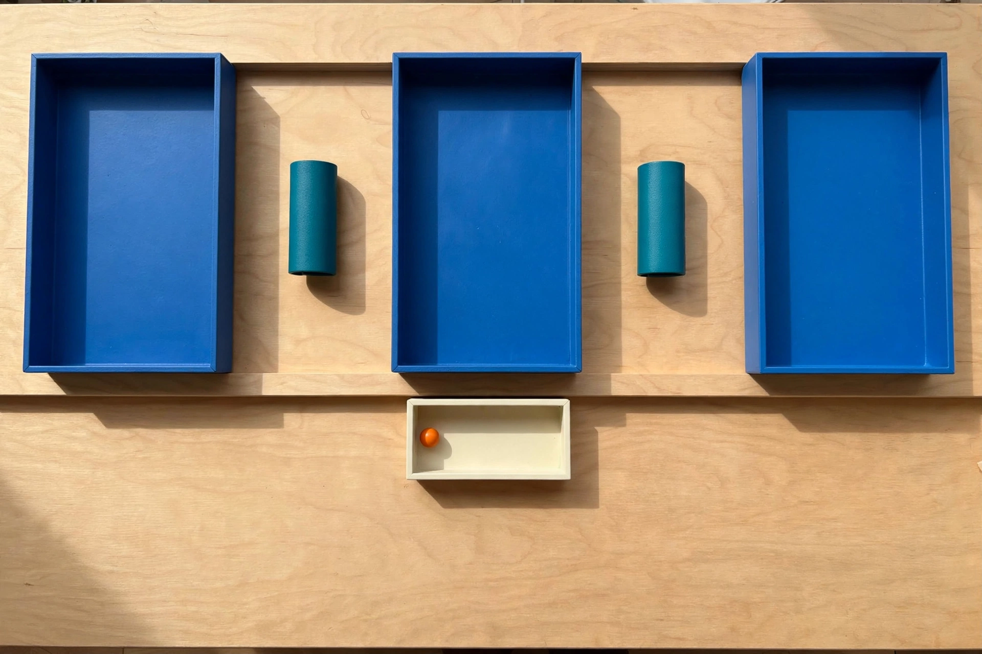 Three blue trays and two teal cylinders are arranged on a wooden surface, with an ivory tray containing an orange bead below.