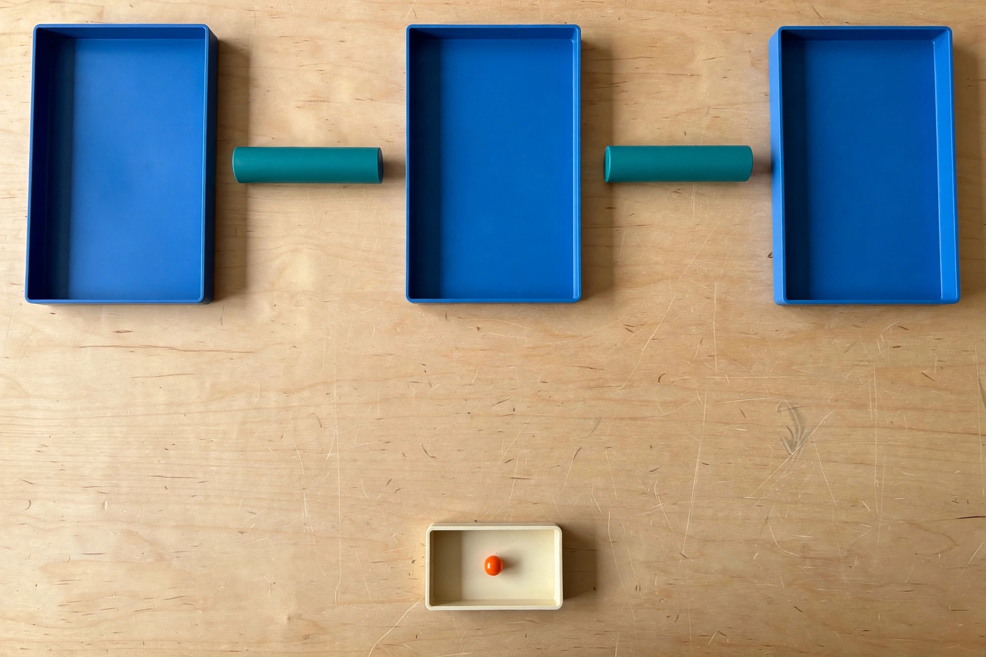 Three blue trays and two teal cylinders are arranged on a wooden surface, with a small ivory tray holding an orange bead below.