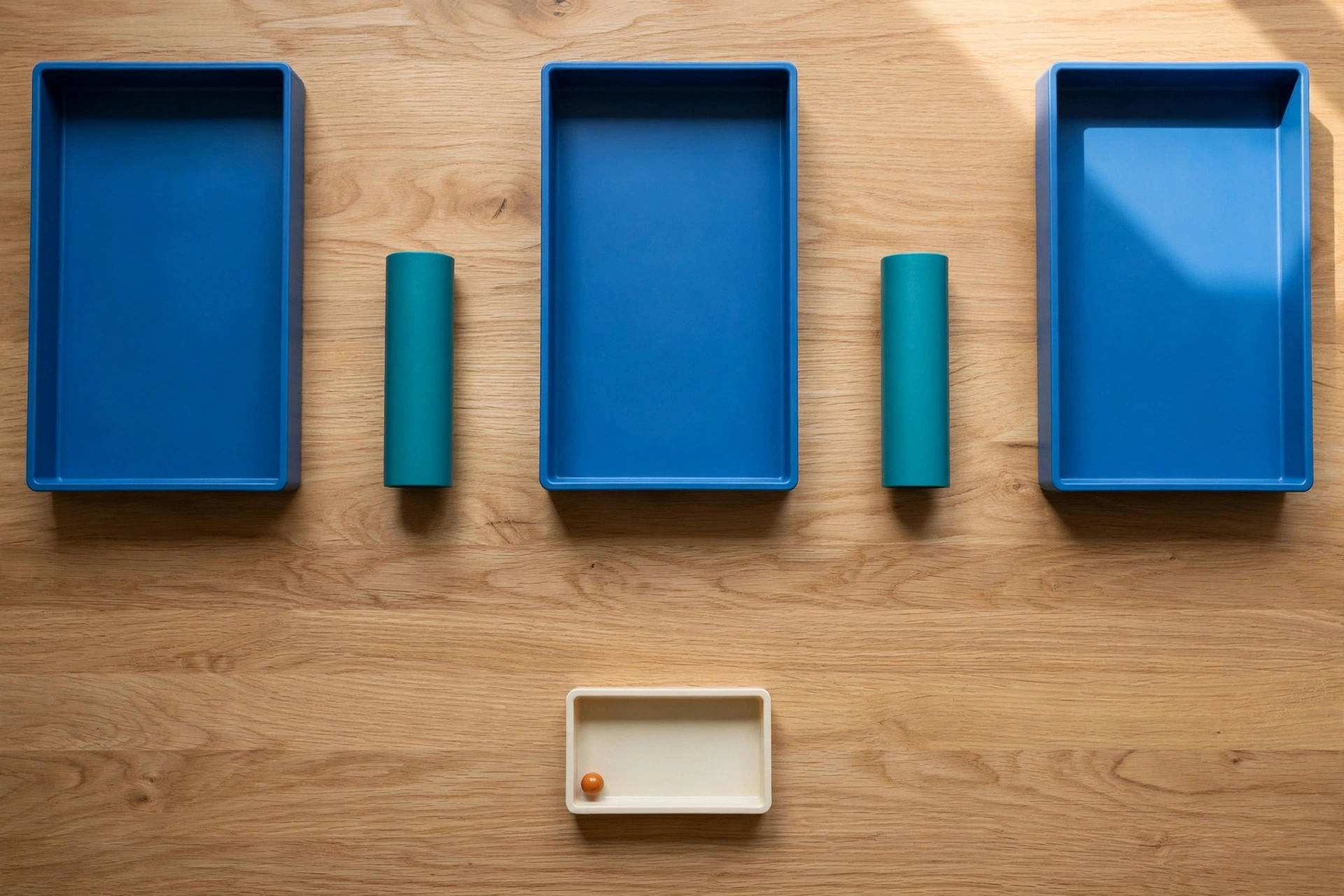 Three blue trays and two teal cylinders are arranged on a wooden desk, with a smaller ivory tray containing an orange bead below.