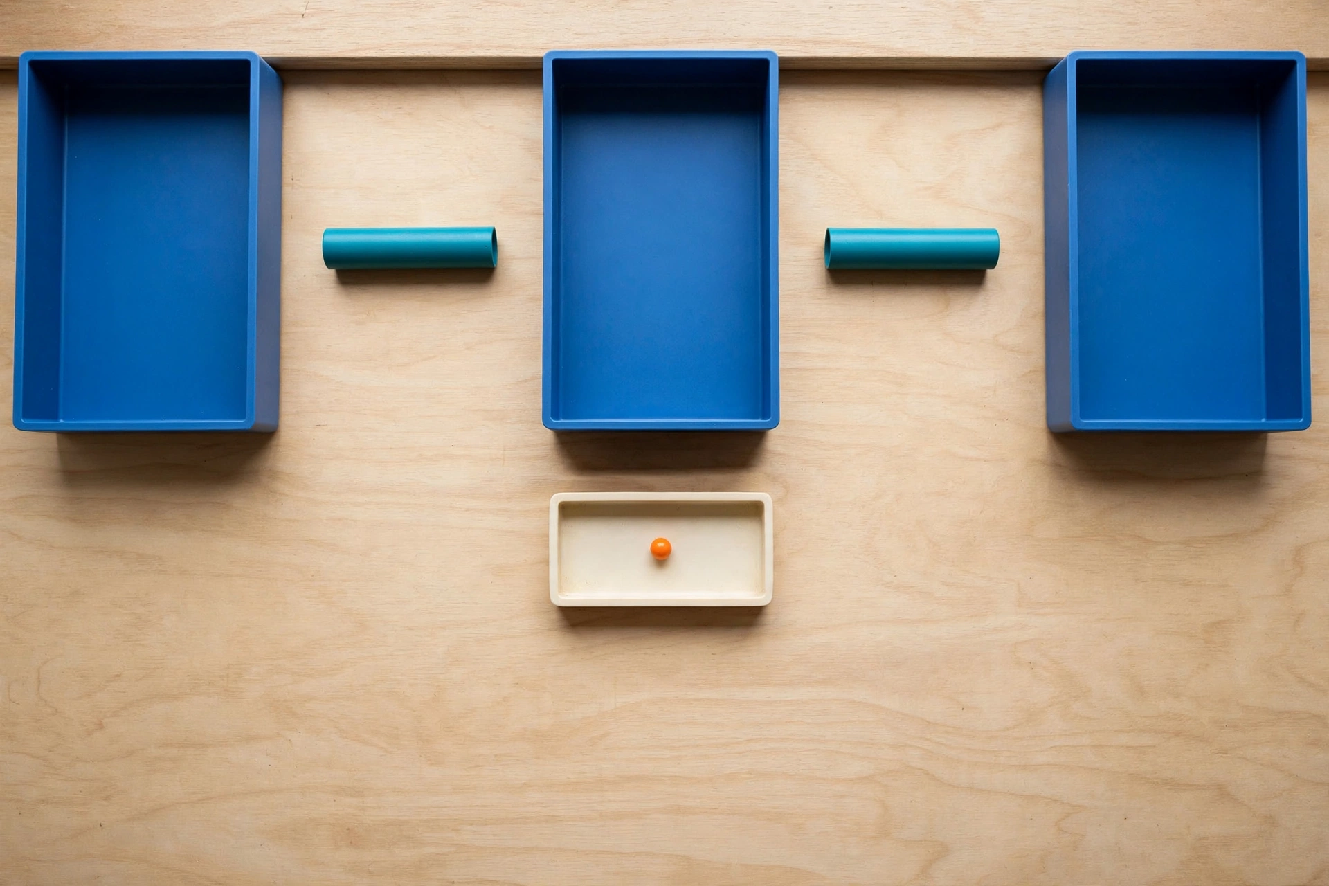 Three blue trays and two teal cylinders are arranged on a wooden surface, with a smaller ivory tray holding an orange bead below.