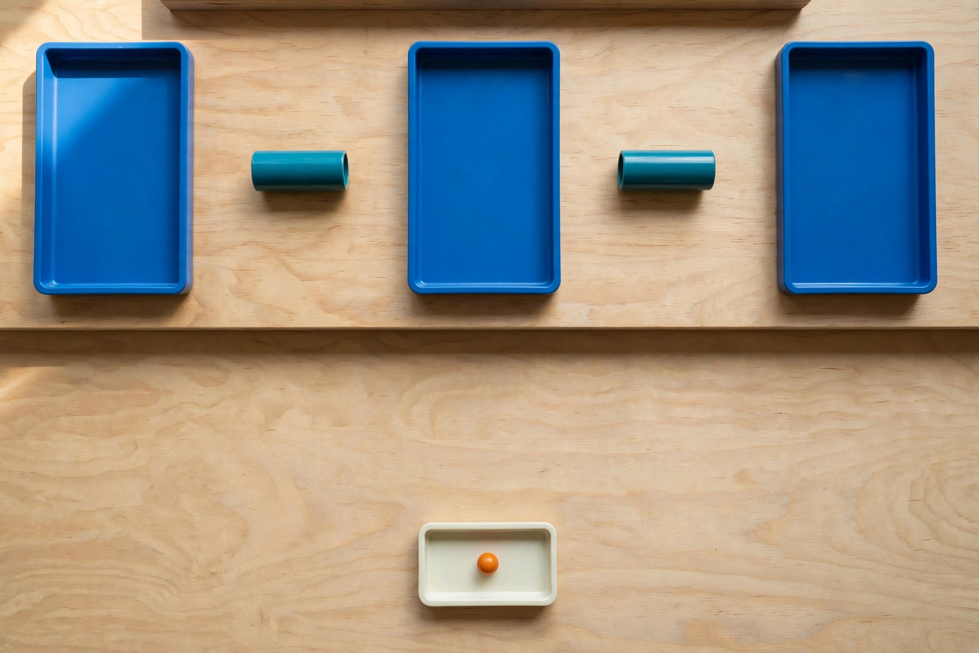 Three blue trays and two teal cylinders are arranged on a wooden surface, with a small ivory tray holding an orange bead below.