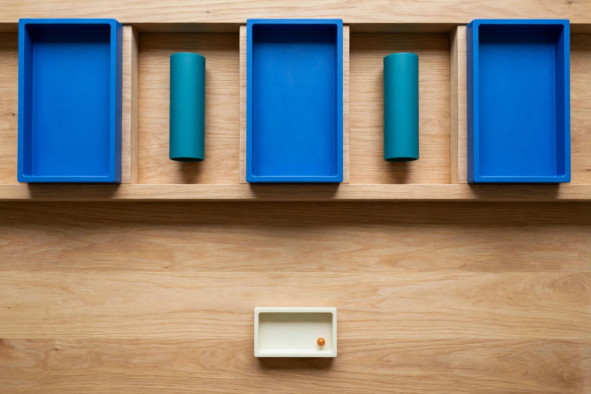 Three blue trays and two teal cylinders are arranged on a wooden surface, with a small ivory tray containing an orange bead below.