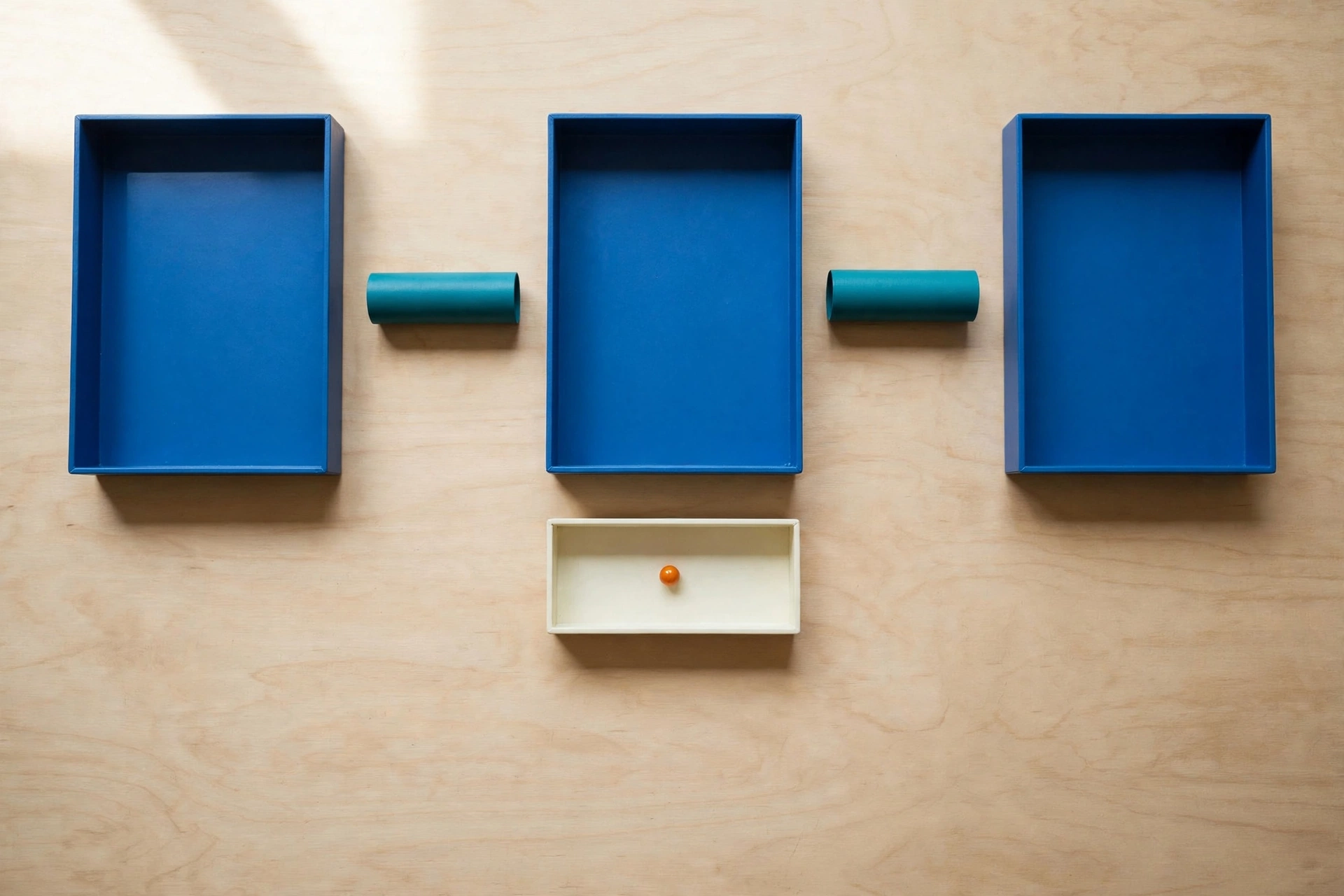 Three blue trays and two teal cylinders are arranged on a wooden surface, with an orange bead in a lower ivory tray.