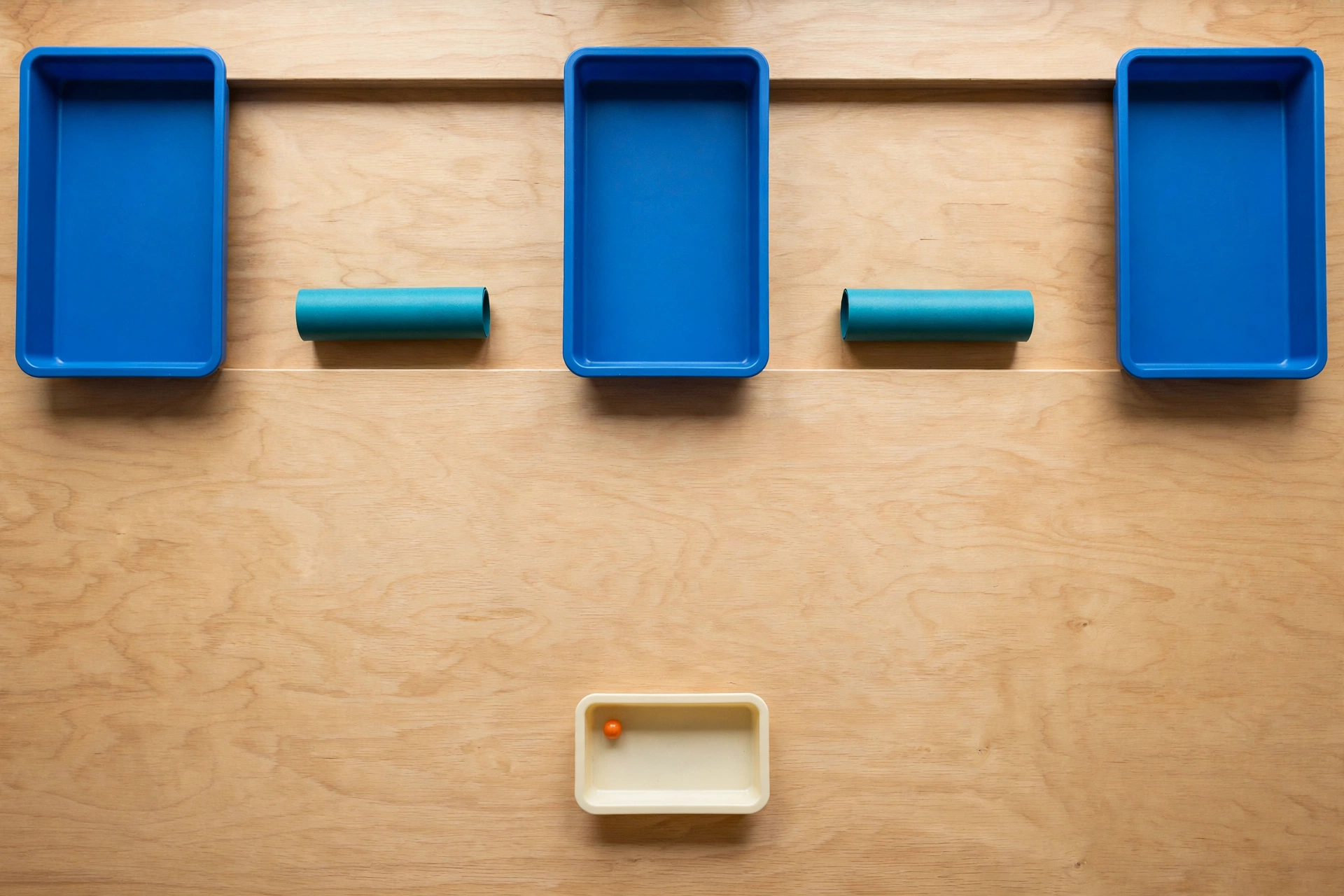 Three blue trays and two teal cylinders are arranged on a wooden surface, with a smaller ivory tray containing an orange bead below.